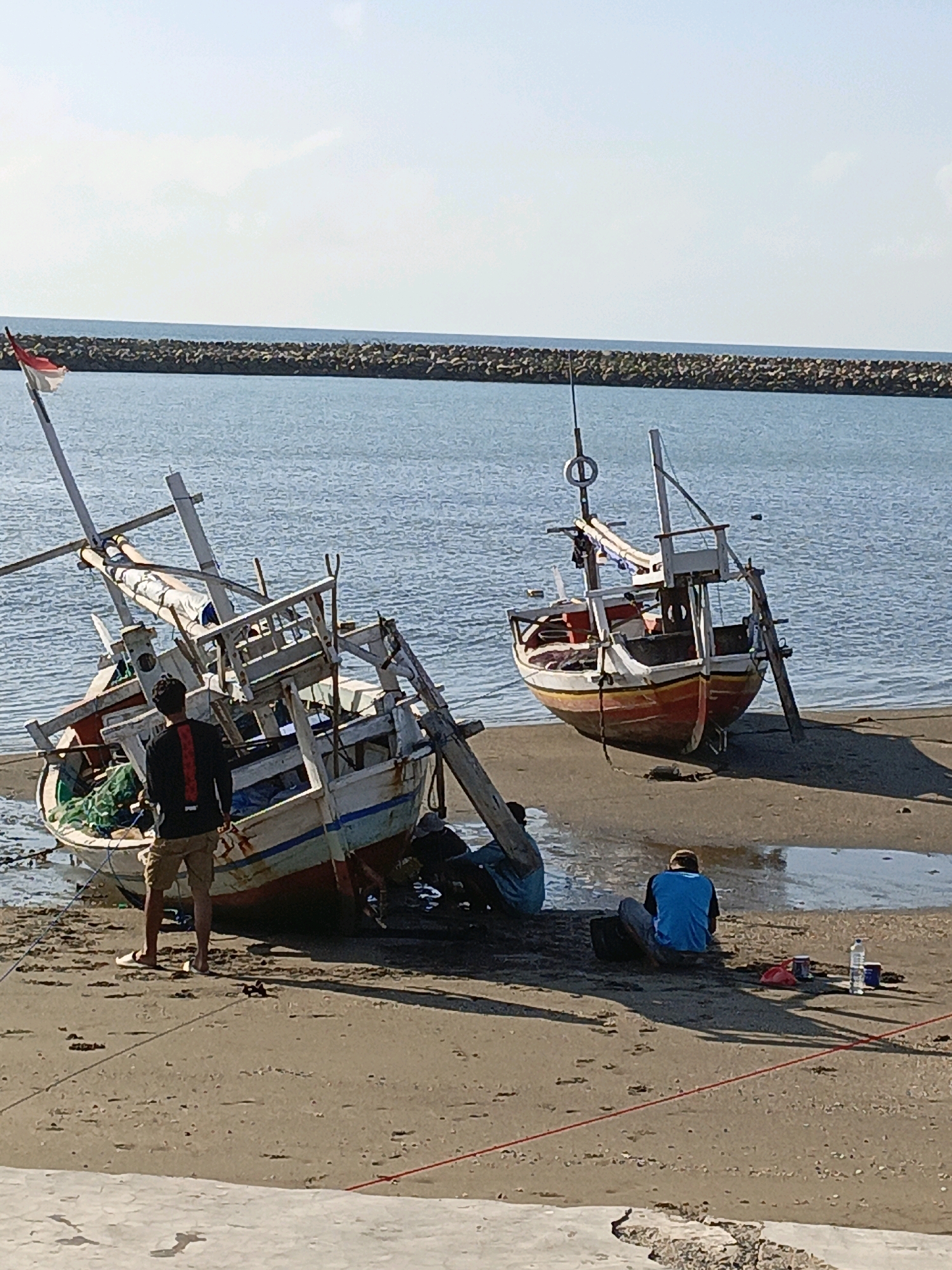 At Ela-ela, Indonesia, two fishing boats are beached on a sandy shore. The boat on the left is tilted and appears to be undergoing repairs, with several individuals visible working on or around it. One person stands near the boat, facing away from the camera. Another boat, also beached, is located to the right of the first. Several people are seated on the sand in the foreground. The water extends to the horizon, with a breakwater made of large rocks visible in the distance under a clear sky.