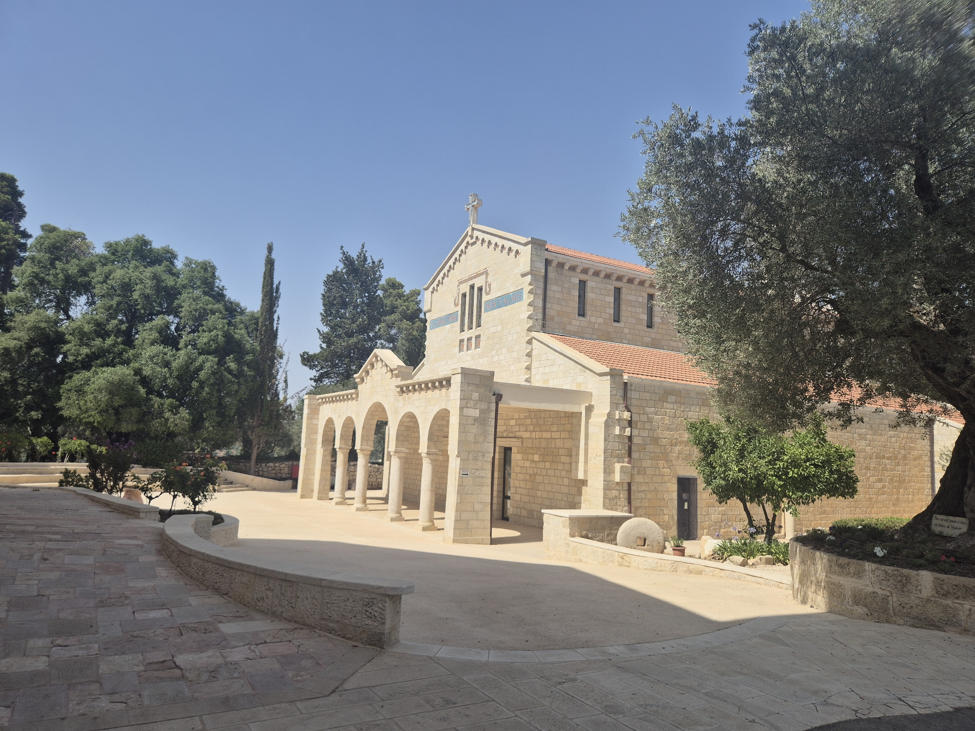 The image captures a view of the Church of the Covenant in Abu Gosh, Israel. The building is constructed from light-colored stone blocks with a red-tiled roof. A prominent feature is a covered walkway with a series of arches supported by columns, leading to an entrance. A cross is visible at the apex of the building's gable.
In the foreground, a paved pathway made of irregular stone slabs curves to the left. Adjacent to the pathway, a low stone wall encloses a landscaped area with a small tree and some flowering plants. To the right of the building, a large, mature olive tree with dense foliage is partially visible. The ground in front of the church appears to be a sandy or gravel area.
The scene is bathed in sunlight, casting shadows that highlight the architectural details. The sky is clear and blue. The overall impression is one of peace and tranquility.