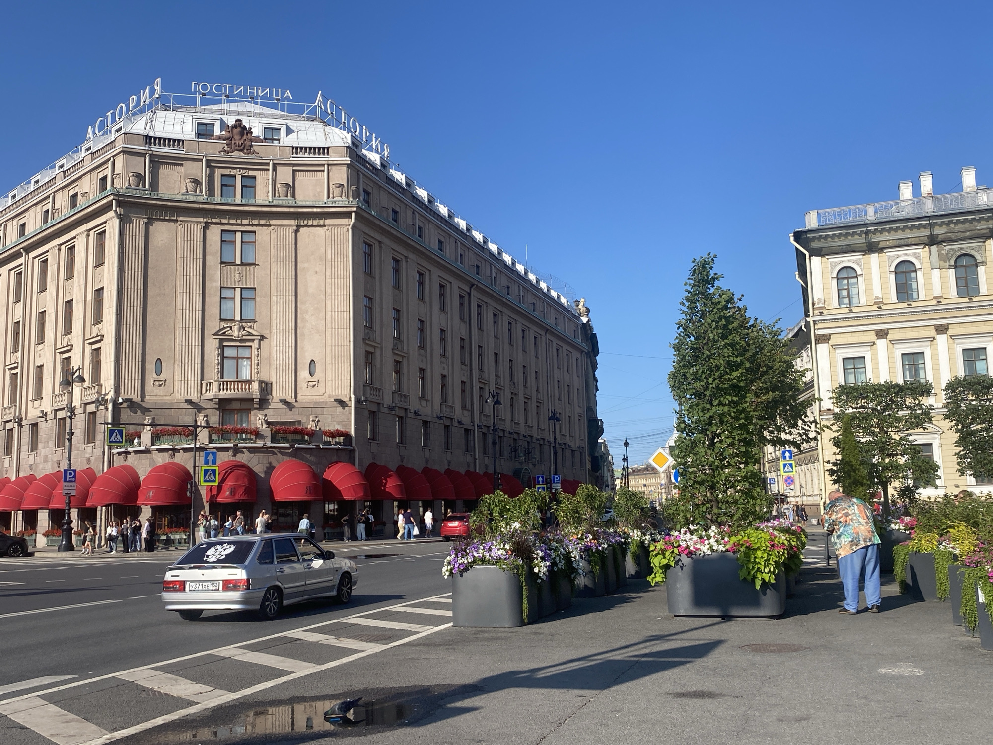 The image depicts a street scene in Saint Petersburg, Russia, featuring the iconic Astoria Hotel. The hotel, a prominent building with classical architecture, dominates the left side of the frame. Its facade is characterized by rows of windows, decorative elements, and a prominent rooftop sign that reads "ГОСТИНИЦА АСТОРИЯ" (Hotel Astoria). Red awnings cover the ground-floor entrances, and several people are seen gathered near the hotel.
In the foreground, a silver sedan is driving on the road, its license plate visible. The road is wet in places, reflecting the bright blue sky. Along the sidewalk, large planters filled with colorful flowers and greenery create a decorative barrier. A man in a vibrantly patterned shirt and blue pants is bending down, tending to the plants.
To the right of the Astoria Hotel, another building with yellow-colored facade and arched windows is visible, suggesting a continuation of the city's urban landscape. The overall scene is bathed in sunlight, indicating a clear day. Pedestrian crossings and street signs are also present, typical of a city environment.