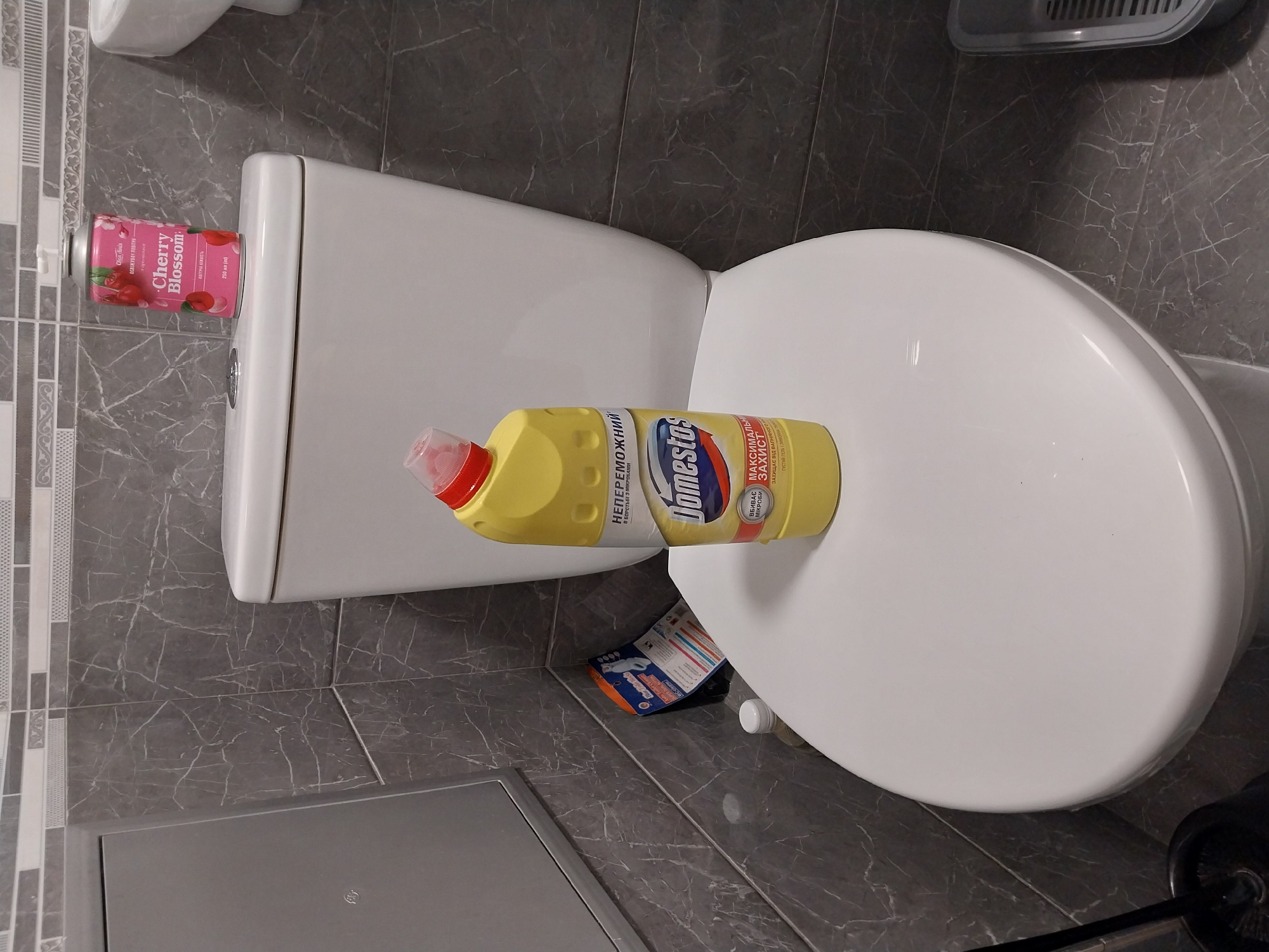 A bright yellow bottle of Domestos toilet cleaner, with a red cap, rests on the lid of a white toilet. The bottle's label, written in Cyrillic, indicates it is "undefeatable in the fight against microbes" and provides "maximum protection against limescale." Next to it, a pink aerosol can labeled "Cherry Blossom Air Freshener" is also placed on the toilet lid. The toilet is situated in a bathroom with grey marble-patterned tiles. A black toilet brush holder is partially visible to the right of the toilet. The lighting suggests it is daytime, and no people are present in the scene.