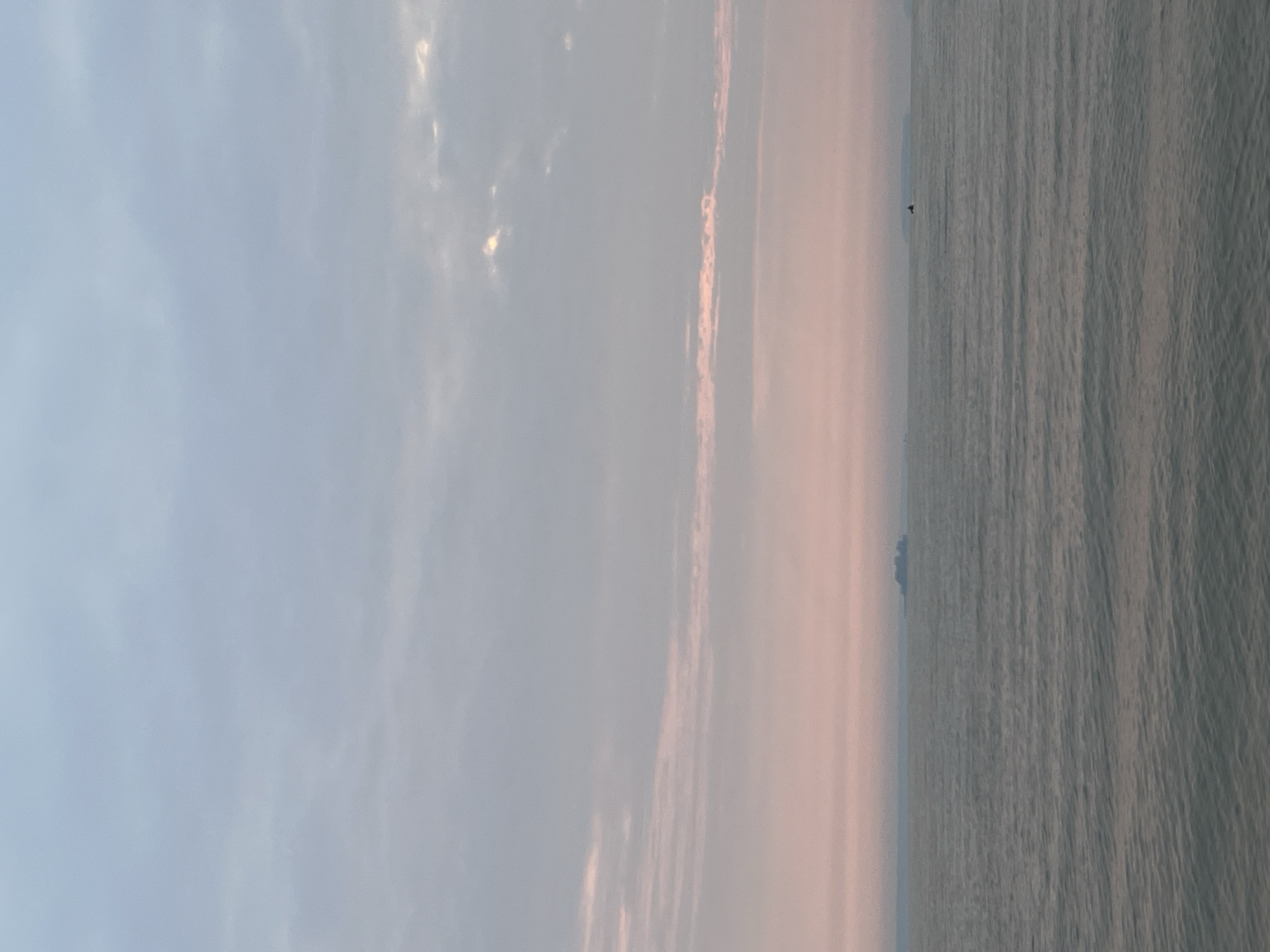 The image shows a wide expanse of grey, rippling water under a pastel sky. The sky transitions from a pale blue at the top to a soft pink near the horizon. Thin, horizontal clouds streak across the sky, some tinged with pink. In the distance, on the horizon, a cluster of dark, indistinct shapes suggests land or a large vessel. Further to the right, a small dark shape, possibly a boat, is visible on the water. The scene is calm and serene. The location is the city of Zav'yalovo, Russia.