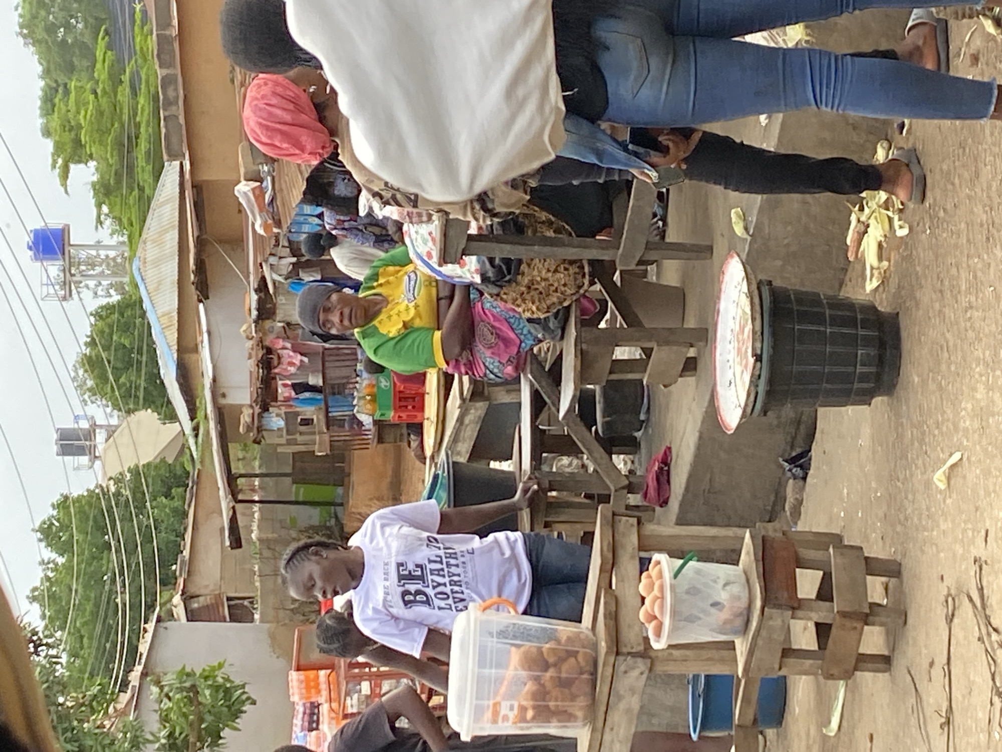 In Bukuru, Nigeria, a bustling outdoor market scene unfolds. In the foreground, a woman in a white t-shirt emblazoned with "BE LOYAL TO EVERYTHING" stands next to a clear plastic tub filled with fried dough balls. Beside her, a small wooden stool supports a white bucket overflowing with eggs. Further back, a woman in a green and yellow shirt and a pink headscarf sits at a wooden table. In the background, market stalls are visible, with various goods and crates of drinks. The environment suggests a warm, possibly humid day with overcast skies. The ground is a mix of dirt and concrete, with scattered debris. Several other individuals are present, their interactions and activities contributing to the lively atmosphere of the market.