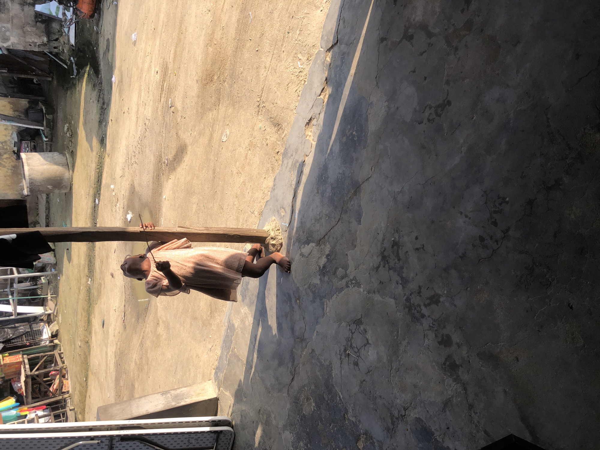 The image depicts a child standing next to a wooden post in a dusty, outdoor environment, likely a street or yard in Ado, Nigeria. The child, seen from the waist down, is wearing a light-colored, possibly peach-colored, dress that reaches below the knees. Their head is tilted back, facing upwards, with their hands appearing to be around the post. The ground in the foreground is a cracked, weathered concrete surface, with shadows cast from the post and an unseen structure. In the background, the ground is a lighter, sandy or dirt material, with scattered debris and puddles of water. A makeshift market stall or collection of structures, constructed from wood and various materials, is visible to the left, with colorful fabrics hanging. The overall lighting suggests it is daytime, with sunlight casting strong shadows. The scene appears to be in an informal or developing urban area, common in parts of Ado, Nigeria.