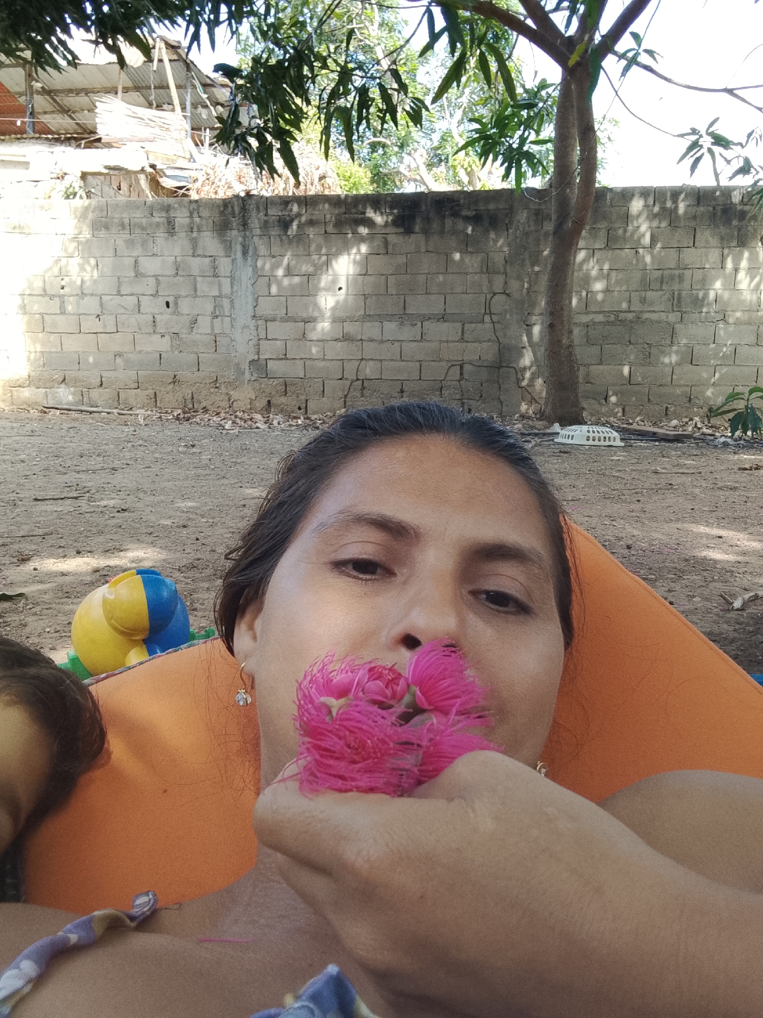 A woman with dark hair is lying down in an outdoor setting, possibly Altagracia, Venezuela. She is holding a cluster of bright pink flowers near her face. Her left eye is visible, and she is wearing gold earrings. She appears to be reclining on an orange surface. A small portion of a child's face is visible in the lower left corner. In the background is a weathered brick wall, a tree, and a partial view of a roof. A yellow and blue plastic object sits on the ground near the wall. The ground appears to be dirt or gravel. The time of day is either dusk or dawn, based on the amount of light.
