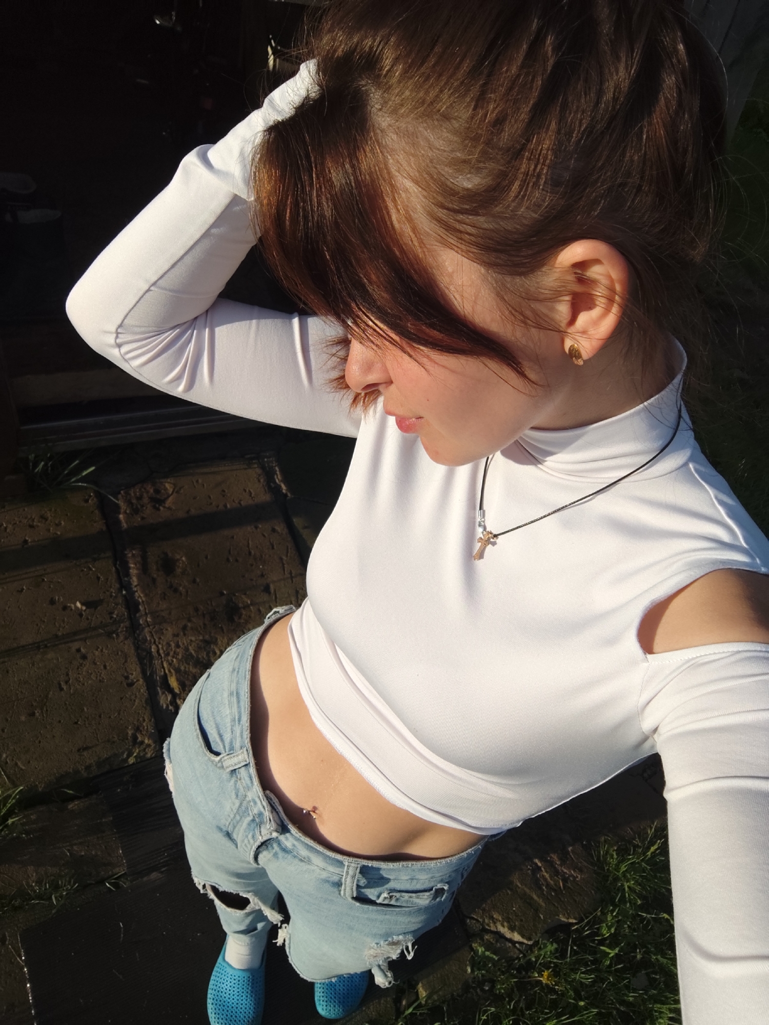 The image captures a young person from a high, slightly overhead perspective in an outdoor setting in Фоки, Russia. The subject, likely female, has brown hair styled up, with some wisps framing her face. She is looking downwards and to her right, with one arm raised, hand near her head, in a casual pose.
She is dressed in a fitted white long-sleeved top featuring a high neck and distinctive cut-out shoulders. Around her neck, she wears a thin black cord necklace adorned with a small gold-toned cross pendant and a silver bead. A small, shiny gold-toned earring is visible in her right ear. Her lower half is clad in light blue, distressed denim jeans, revealing her midriff and a belly button piercing with a small stud. She wears white socks and bright blue, textured slip-on shoes.
The scene depicts an outdoor space, possibly a backyard or patio. The ground beneath her is a mix of dark, irregular paving stones or flagstones and sparse patches of green grass, some appearing damp. The background is predominantly dark and shadowed, suggesting a shaded area or structure. The lighting is characterized by strong, warm, low-angle sunlight, indicative of either late afternoon or early morning, casting long and defined shadows. No visible text is present in the image. The overall mood is casual and contemplative.