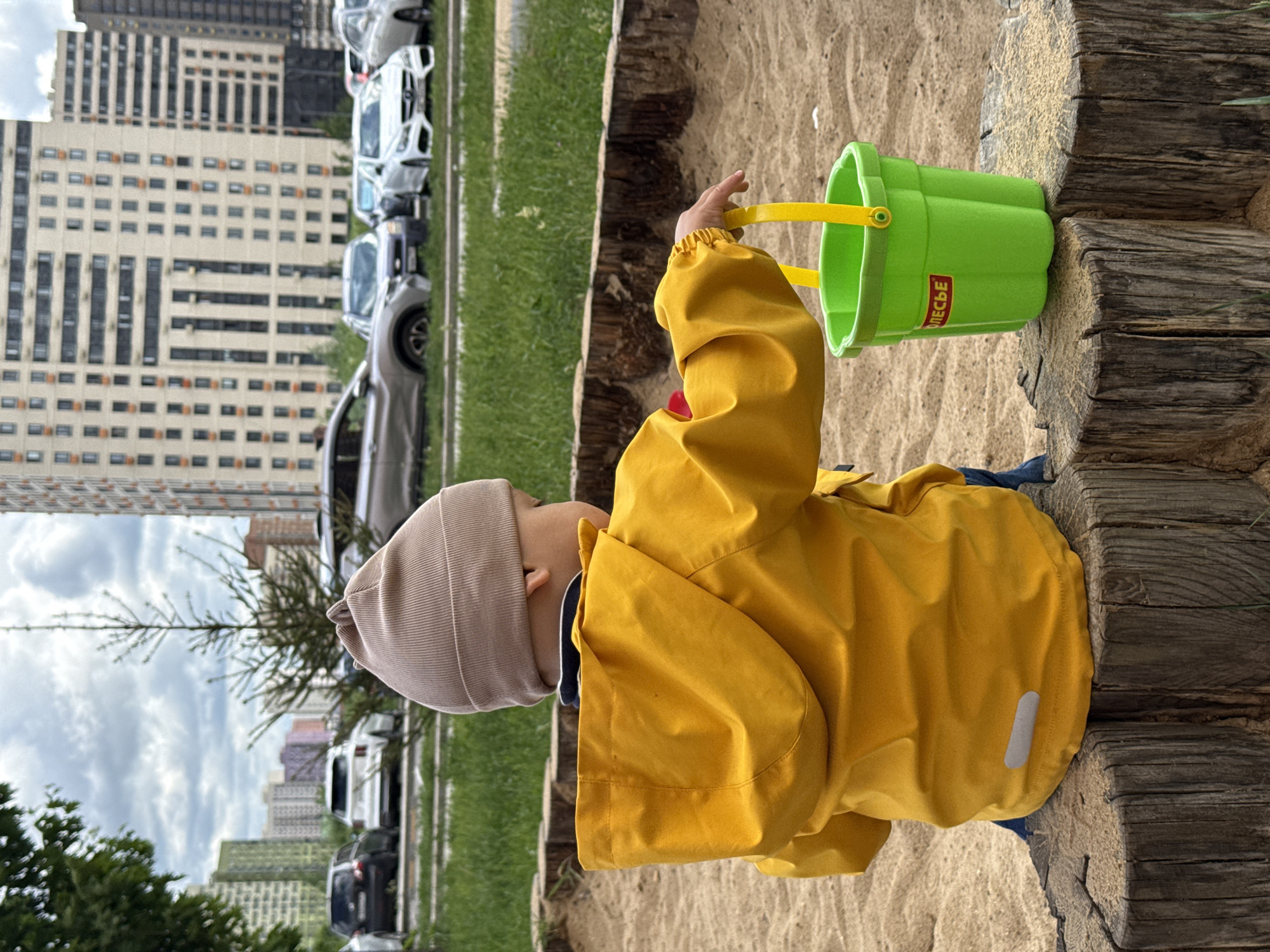 A young child in a yellow jacket and beige hat is playing in a sandpit with a green bucket. The bucket has a yellow handle and the word "ПОЛЕСЬЕ" is printed on the side. The sandpit is bordered by weathered wooden logs.
Behind the sandpit, there is a strip of green grass, followed by a road with parked cars. In the background, there are tall apartment buildings typical of urban Moscow. The cloudy sky suggests the weather is overcast. The buildings indicate the scene is likely within the city limits of Moscow, Russia.