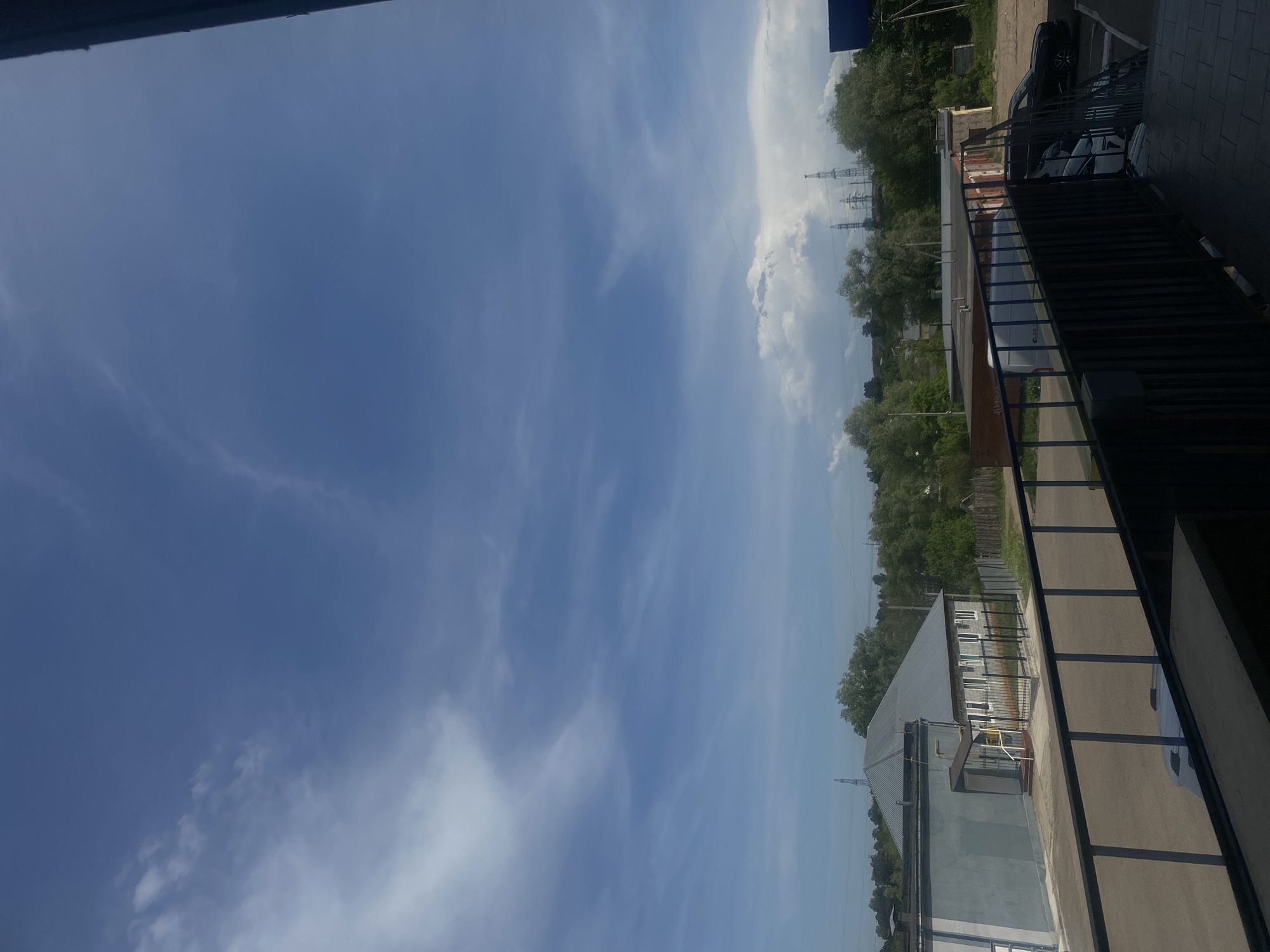 The image shows a view from an elevated position, possibly a balcony or upper floor of a building, in what appears to be a suburban or industrial area of Chaikovsky, Russia. The sky dominates the left side of the image, exhibiting a clear blue color interspersed with wispy cirrus clouds.
In the midground, a row of trees lines the horizon, partially obscuring some industrial structures, including power pylons, that are set back. The foreground includes part of a building with a corrugated metal roof, and what appears to be a parking lot. A black metal railing runs along the edge of the balcony. A black car is visible in the parking lot.