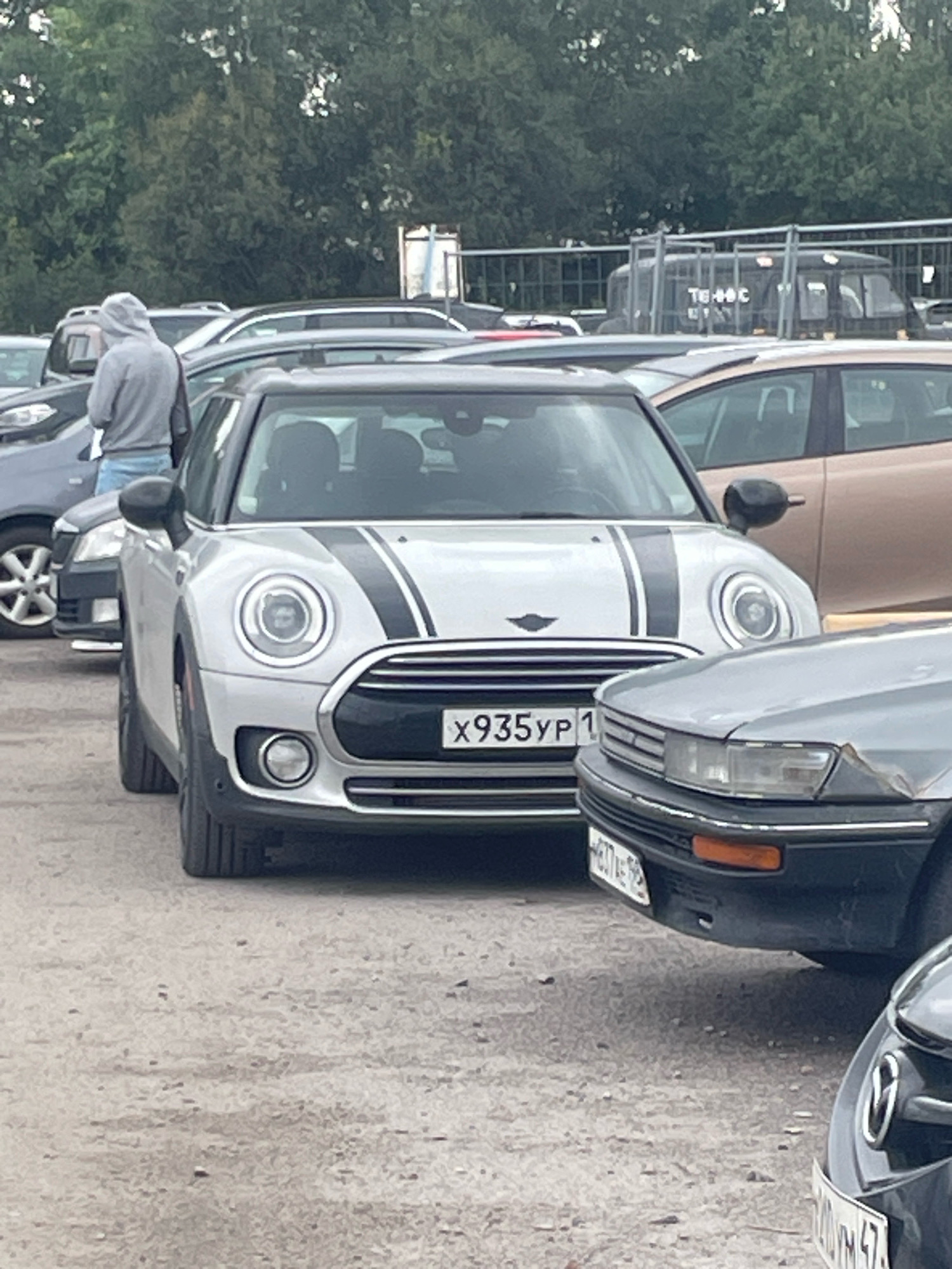 A light silver Mini Cooper is parked on a gravel lot with trees and a fence in the background. A person in a grey hoodie and jeans is standing next to the car on the driver's side. The Mini Cooper has two black racing stripes on the hood and a license plate that reads "X935YP 1". A dark gray car is parked to the right of the Mini Cooper, and the front of a black car is visible in the foreground. The image was taken in the city of Saint Petersburg, Russia.