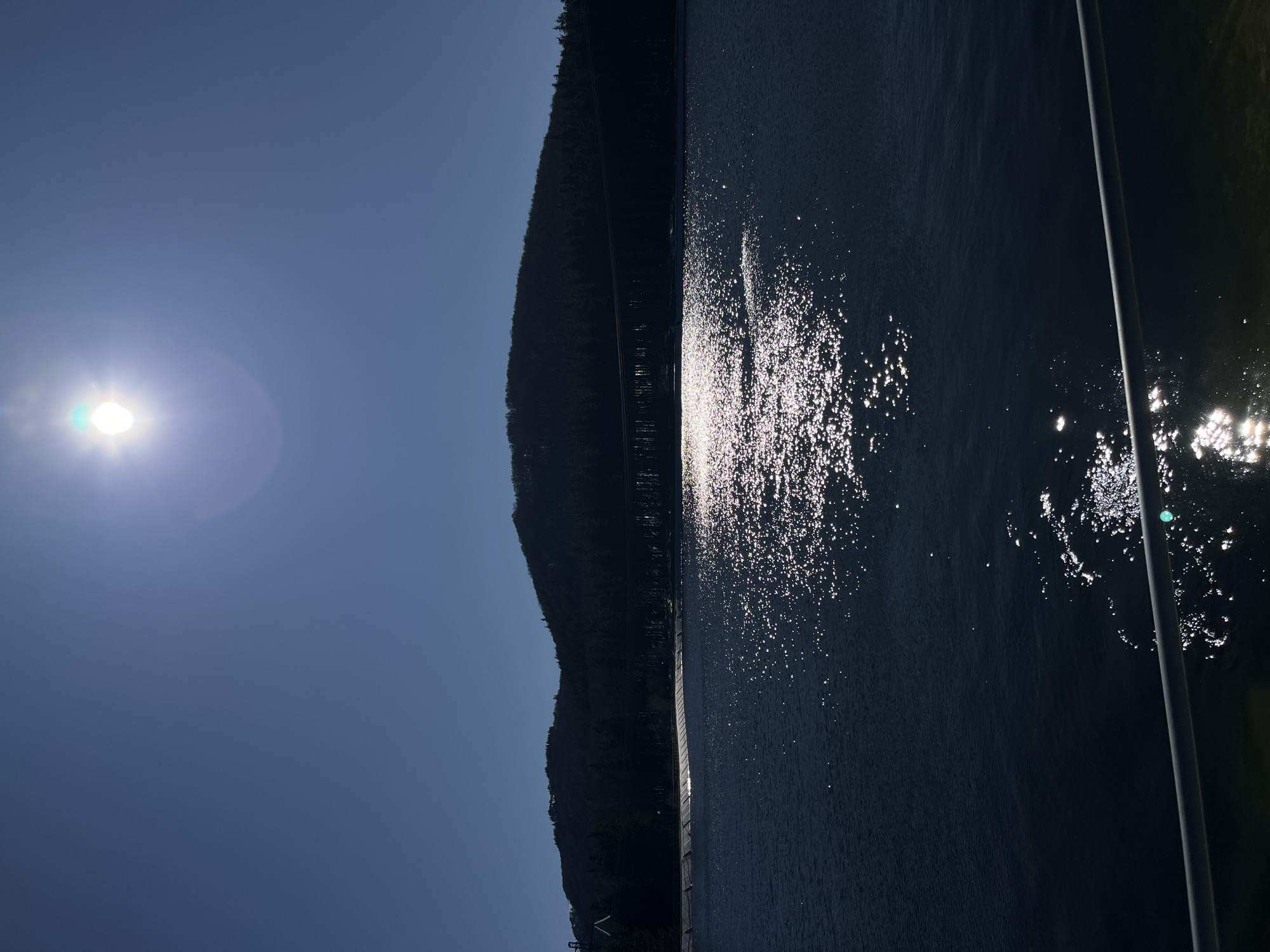 The image shows a body of water reflecting sunlight. A dark, tree-covered mountain is visible on the left side of the frame. A vertical pole is present on the right side of the image. The sky is clear and blue, with the sun positioned in the upper left corner. The location in the city of Barziya, Bulgaria is not known without additional information.