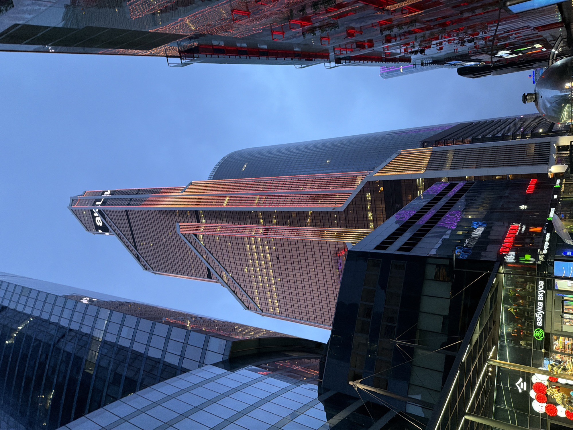 This media file captures a modern urban landscape at dusk or early evening, likely in Khimki, Russia, as indicated by context. The scene is dominated by several contemporary high-rise buildings constructed primarily of glass and steel. The sky is a clear, deep blue, transitioning into twilight.
In the center, a prominent skyscraper features distinctive vertical strips of red and orange lights illuminating its facade, creating a striking visual contrast against the darkening sky. Its glass surfaces reflect the ambient light and parts of other structures. To the left, another large glass building reflects the sky and surroundings in its gridded panels.
On the right side, at the base of a dark, reflective building, are several brightly lit storefronts. A prominent sign in Cyrillic clearly reads "азбука вкуса" (Azbuka Vkusa), which is a well-known Russian supermarket chain. Other partial or less distinct Cyrillic text is visible on nearby signs and displays.
The scene is static, with no people or overt activity visible, emphasizing the architectural elements and the play of artificial lights in a modern city environment. The overall impression is one of a vibrant commercial or business district.