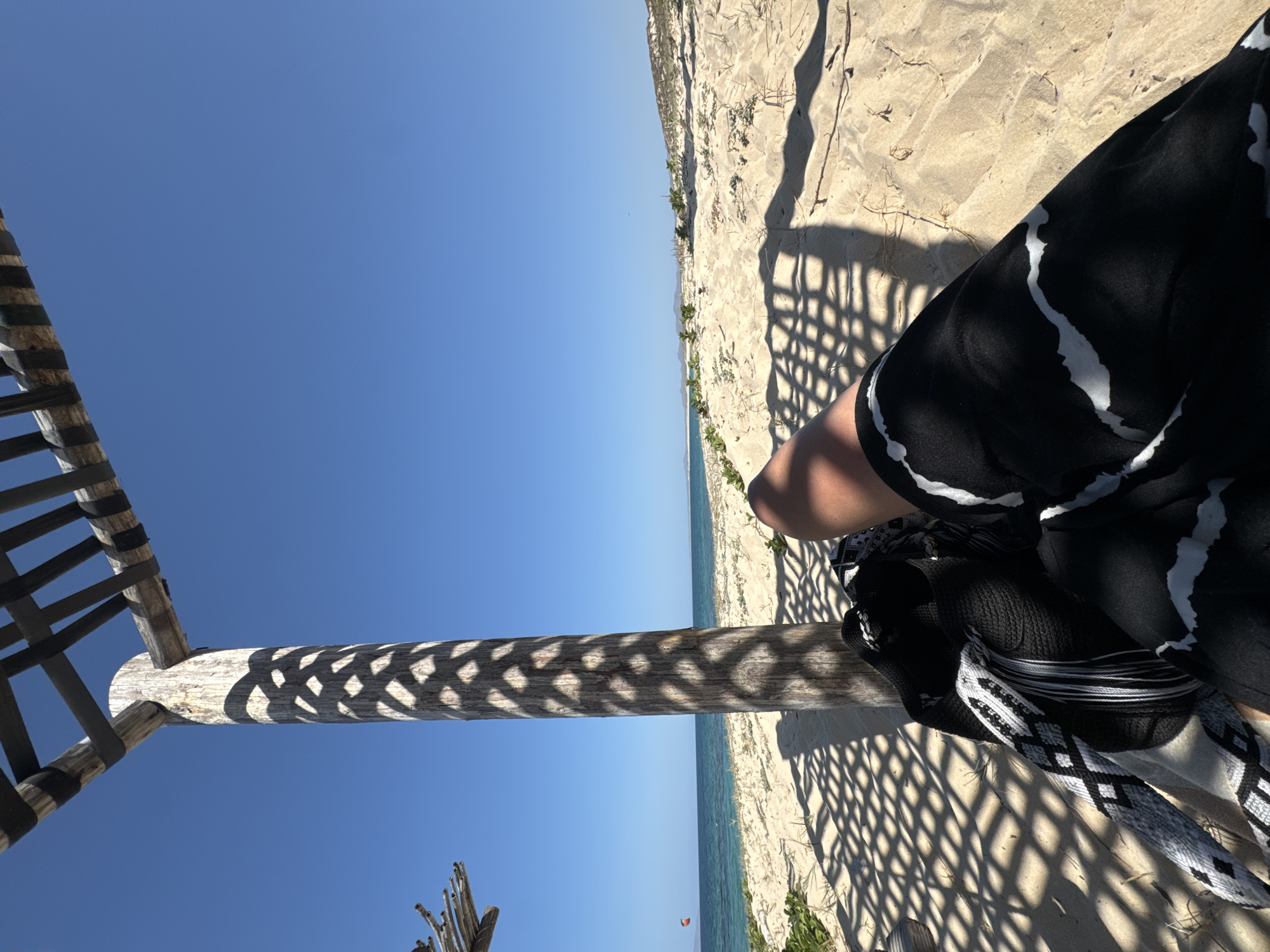 The image shows a beach scene taken from under a shaded structure, likely a palapa or similar beach shelter. A wooden post supporting the structure runs vertically through the center of the image, covered in a patterned shadow. To the left of the post is a clear blue sky. To the right of the post is a sandy beach, meeting the ocean in the distance. Part of a person is visible in the lower right, wearing a dark garment with white patterns. A black woven bag with a white strap is also visible. Vegetation grows along the edge of the beach. The exact location within the city of Unknown, Unknown is not discernible from the image.