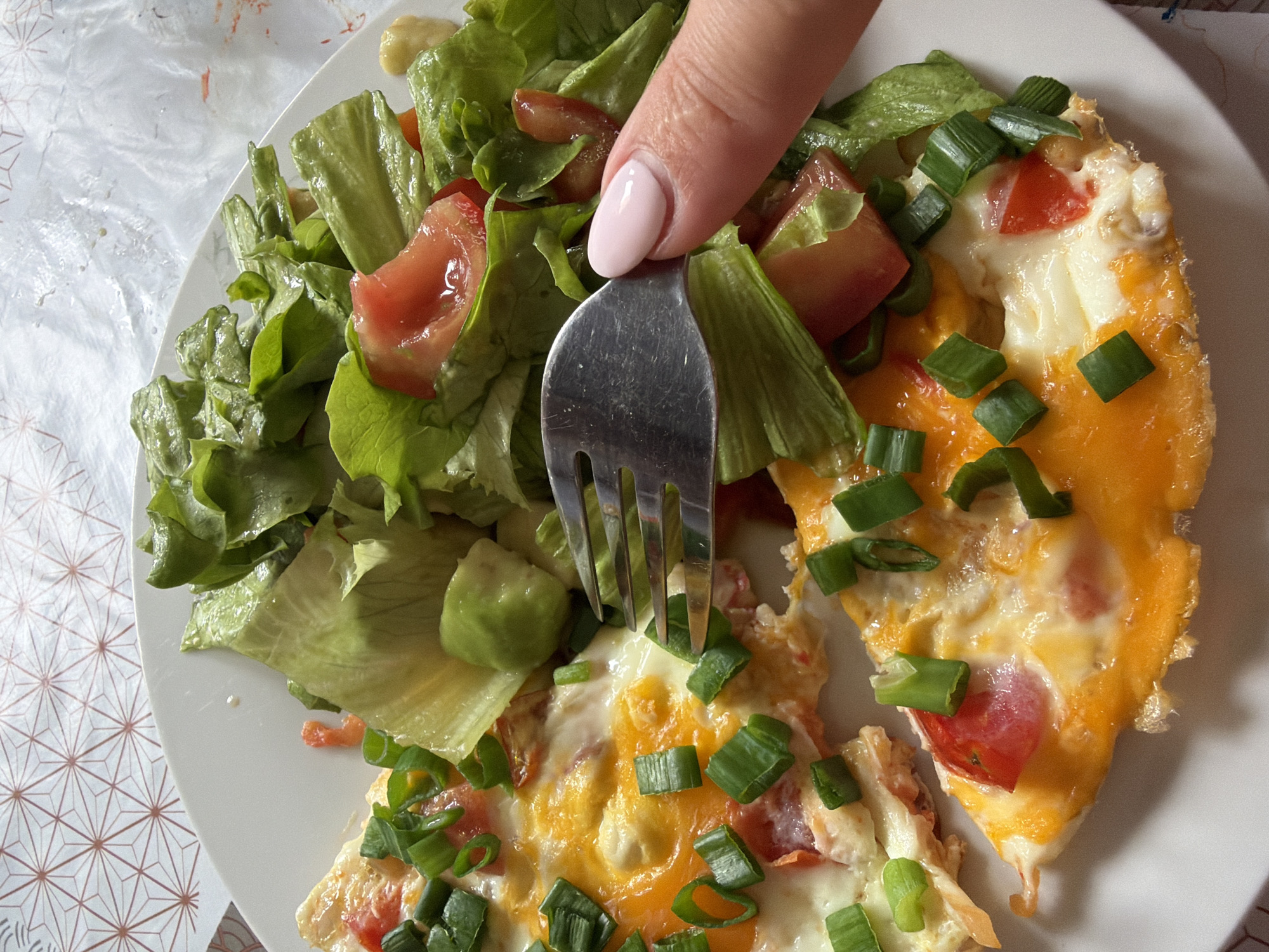 A top-down, close-up shot captures a white plate filled with food. On the left side of the plate, there's a salad composed of lettuce and tomato slices. Adjacent to the salad, two pieces of omelet are visible, topped with chopped green onions and tomato pieces. A hand with a pale pink nail polish is holding a metal fork, which is inserted into the food. The plate sits on a surface covered with a patterned cloth. The location is not specified in the image and impossible to tell.
