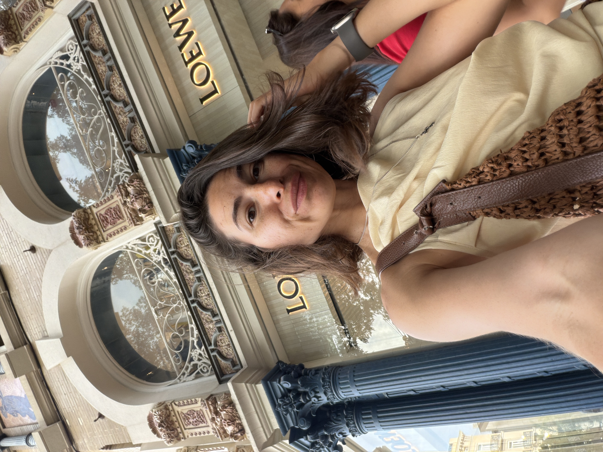 A woman is taking a selfie in front of a Loewe store. She has brown hair and is wearing a cream-colored top and a brown woven purse. The store is ornate with large windows and decorative architectural elements. Another woman with dark hair wearing a red shirt is partially visible in the background to the right. The scene appears to be outdoors in a city environment.