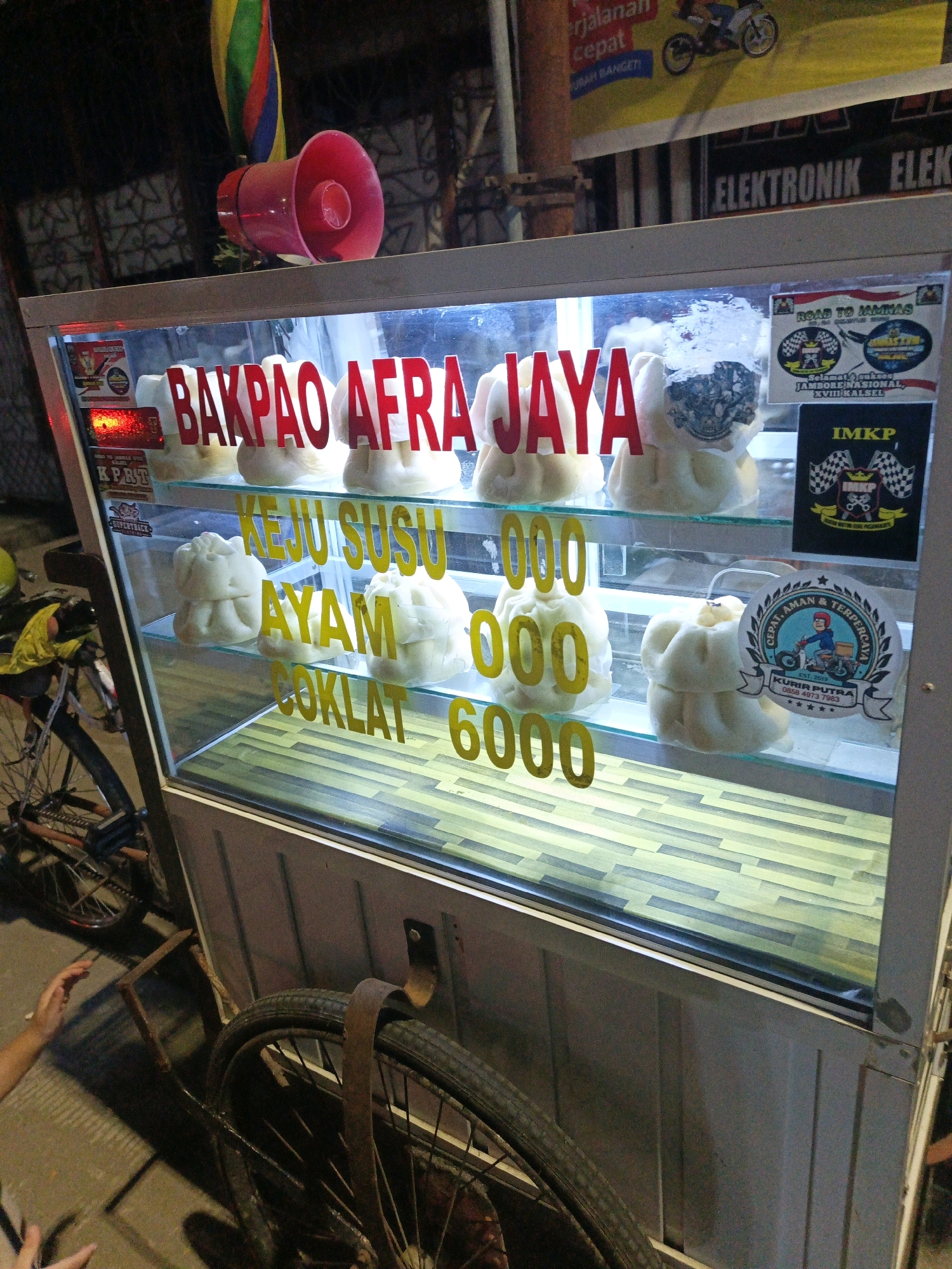 The image shows a food cart selling "Bakpao Afra Jaya" in Pangkajene, Indonesia. The cart has a glass display case filled with steamed buns, which appear to be labeled with flavors such as "Keju Susu" (Cheese Milk), "Ayam" (Chicken), and "Coklat" (Chocolate). There is also text on the cart indicating prices, with "6000" visible. Several stickers are affixed to the inside of the glass display, including one for "IMKP" and another with the text "CEPAT, AMAN & TERPERCAYA" and "KURIR PUTRA". In the background, a street scene is visible with other businesses and what appears to be a motorcycle. A bicycle is parked next to the cart, and a person's hand is visible reaching towards the cart. The lighting suggests it is evening or nighttime.