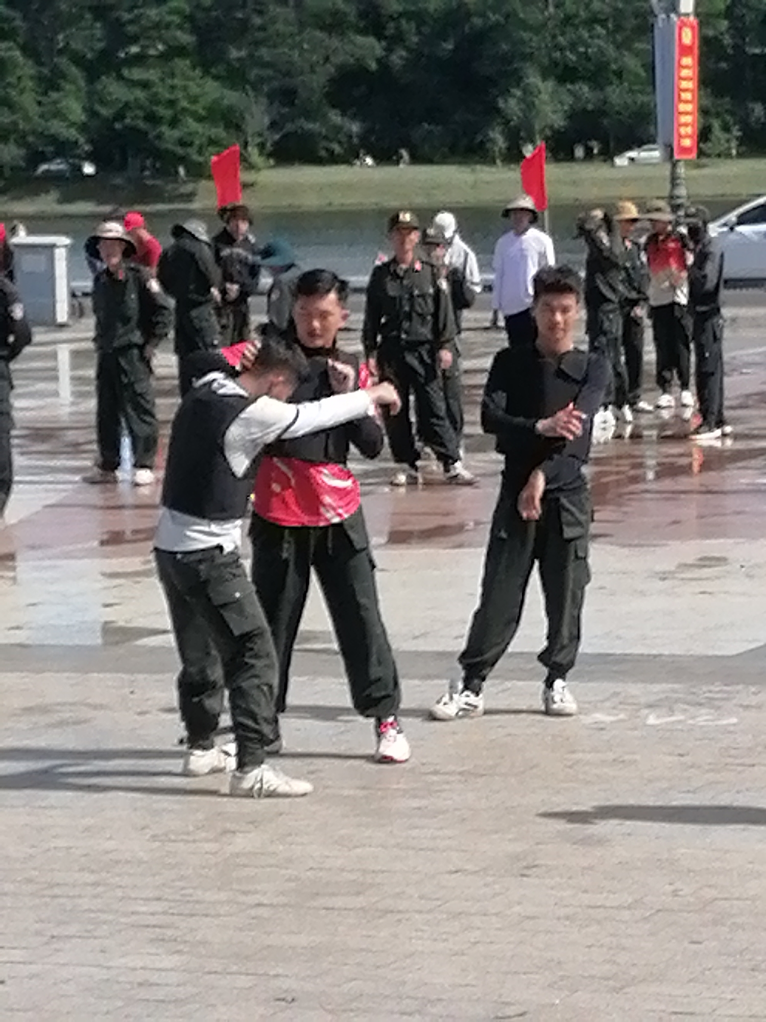 In an outdoor urban setting, potentially a plaza or wide walkway, a group of individuals, mostly men, are engaged in what appears to be a martial arts demonstration or training session. The individuals are dressed in dark, matching uniforms, consisting of long-sleeved shirts and cargo pants, with some wearing vests over their shirts. The ground is a light-colored, textured pavement, with scattered puddles or wet patches suggesting recent rain or water features.
In the foreground, two men are actively interacting. One man, wearing a black vest over a white long-sleeved shirt, is in a defensive posture, with his arms raised and body leaning forward. The other man, dressed in a black long-sleeved shirt and vest, is in an offensive stance, his arms extended and positioned as if to strike or grapple. Both men are wearing athletic shoes.
Behind them, several other individuals in similar uniforms are standing or observing. Some are looking towards the central action, while others are facing away. In the background, more people are visible, some wearing hats and casual attire, suggesting they are spectators or part of a larger event. A body of water, possibly a lake or river, is visible beyond the pavement, with trees lining the far bank. Red flags are present, one prominently displayed on a pole. The presence of a white car in the background suggests a public area with vehicle access. The overall atmosphere appears to be that of an organized event or practice session taking place in a public, possibly scenic, location. The description does not confirm the location as Da Lat, Vietnam.