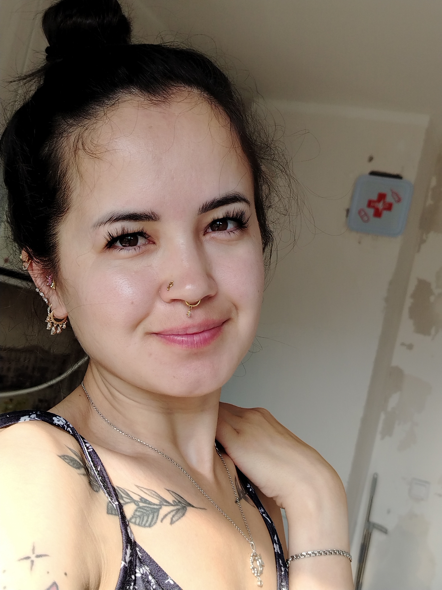 A medium close-up selfie of a woman with her hair in a bun, facing the camera with a slight smile. She has dark hair, prominent eyelashes, and several piercings, including a septum ring and earrings. She is wearing a dark patterned top and a silver necklace. Tattoos are visible on her chest and shoulder. The background appears to be an interior space with a wall that has some peeling paint and a small first-aid kit mounted on it. The lighting suggests it is daytime. The location is not determinable from the image.