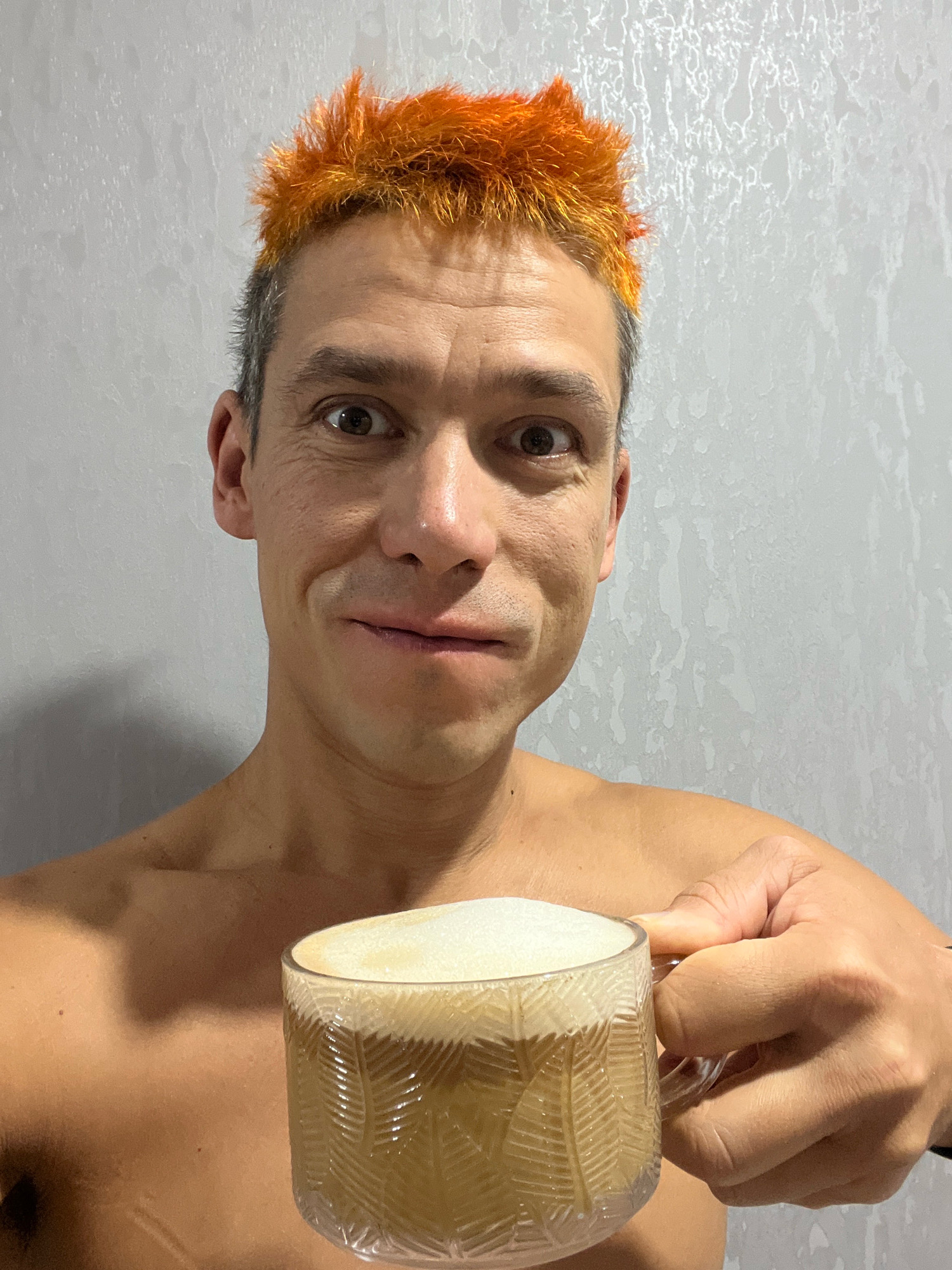 A man with spiky, bright orange hair and a greyish complexion is holding a clear glass mug filled with a frothy, light brown beverage. He is shirtless, revealing his shoulders and upper chest, and his skin appears tanned. The man has dark eyes, a slightly crooked nose, and a subtle, somewhat awkward smile. The background is a textured, light grey wall, possibly a bathroom or shower area. The image is a close-up, focusing on the man's face and the mug he is holding. There are no discernible features that would indicate this scene is located in the city of Chaikovsky, Russia.