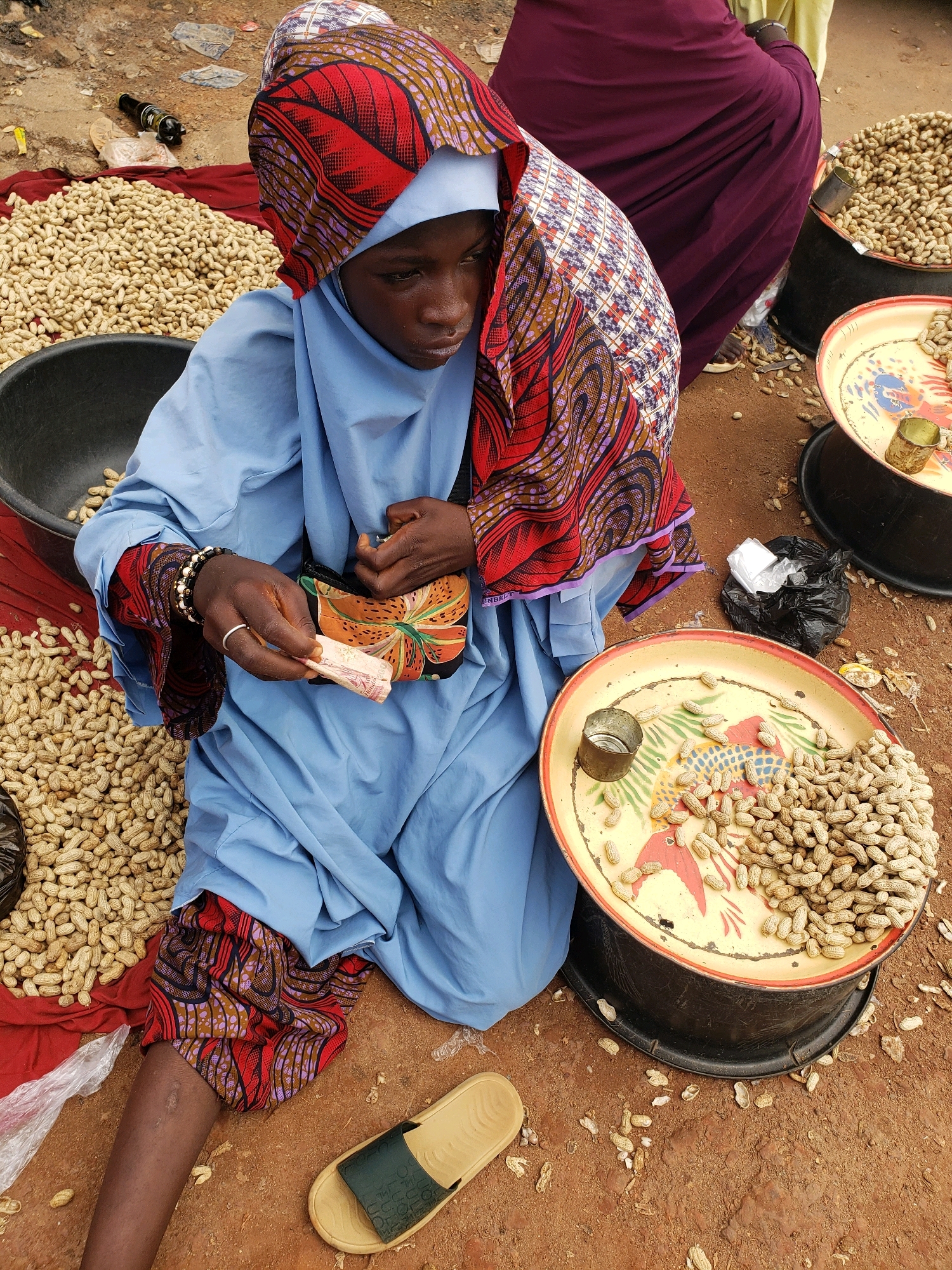 In the city of Sabon Tasha, Nigeria, a young female vendor is seen kneeling on the ground. She is dressed in a light blue tunic and pants with a vibrant, patterned headscarf. Her hands are clasped together, holding a small stack of Nigerian Naira currency. She wears a silver ring on her left ring finger and a beaded bracelet on her left wrist. In front of her, on the ground, is a black bowl filled with peanuts, and a circular tray also filled with peanuts, decorated with colorful bird imagery. A small metal cup sits on the tray. To her right, a discarded black plastic bag lies on the reddish-brown soil, scattered with more peanuts. A single light-colored sandal with a dark green strap rests to her lower right. The vendor appears to be engaged in commerce, with peanuts prominently displayed for sale. The background shows a pile of peanuts on a red cloth and a dark cylindrical object that might be a container or a stall support.