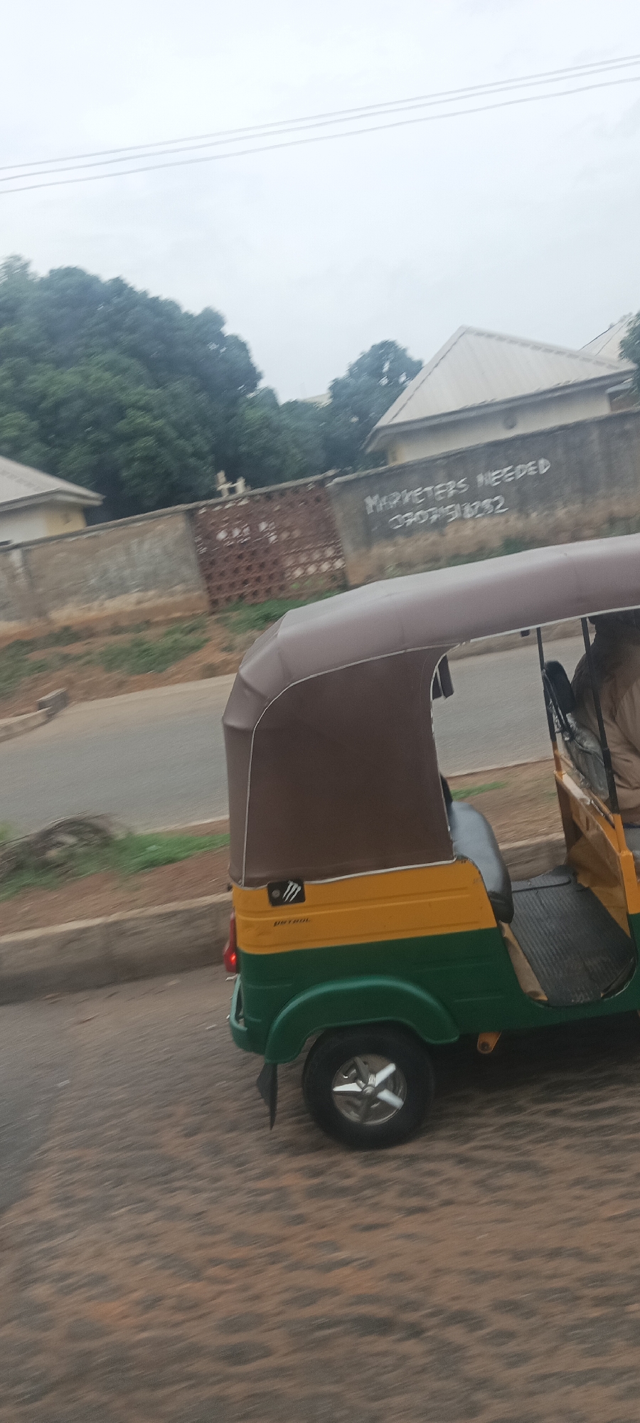 The image captures a side view of a green and yellow Keke Napep (auto-rickshaw) in motion on a road. The vehicle has a brown canopy and a visible "Monster Energy" logo sticker, along with the word "PETROL" written below it. A person is partially visible inside the Keke Napep, likely a driver or passenger. The road surface shows motion blur, indicating the vehicle is moving.
In the background, a concrete wall separates the road from some buildings and dense green trees. On the wall, white spray paint spells out "MARKETERS NEEDED" followed by a phone number, "0907518232". Behind the wall, parts of buildings with light-colored, possibly metallic, roofs are visible amidst the foliage. The sky is overcast and appears hazy or cloudy, suggesting a cloudy day. Overhead, three thin lines, likely power lines, stretch across the sky. The overall scene suggests an urban or semi-urban environment in a tropical or subtropical region, strongly indicative of Nigeria given the phone number format and commonality of Keke Napeps.