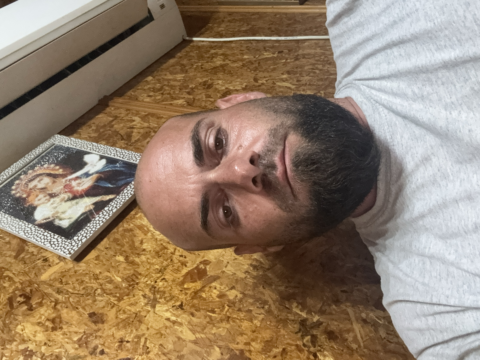 A bald, bearded adult male is depicted in a close-up, front-facing image, presumably in Sarnevo, Bulgaria. He has dark eyes, dark eyebrows, and a full dark beard, with a neutral to serious facial expression. His skin appears slightly shiny. He is wearing a light-colored, possibly heather grey, short-sleeved t-shirt. The background consists of a wall constructed from oriented strand board (OSB), displaying a textured wood-chip pattern in shades of brown and yellow. Mounted on the upper left portion of the wall, behind the man's head, is a framed picture with a white, patterned border, depicting what appears to be a classical religious image. In the upper right corner of the background, a white indoor air conditioning unit is affixed to the wall. A white electrical conduit or cable runs vertically down the right side of the OSB wall.