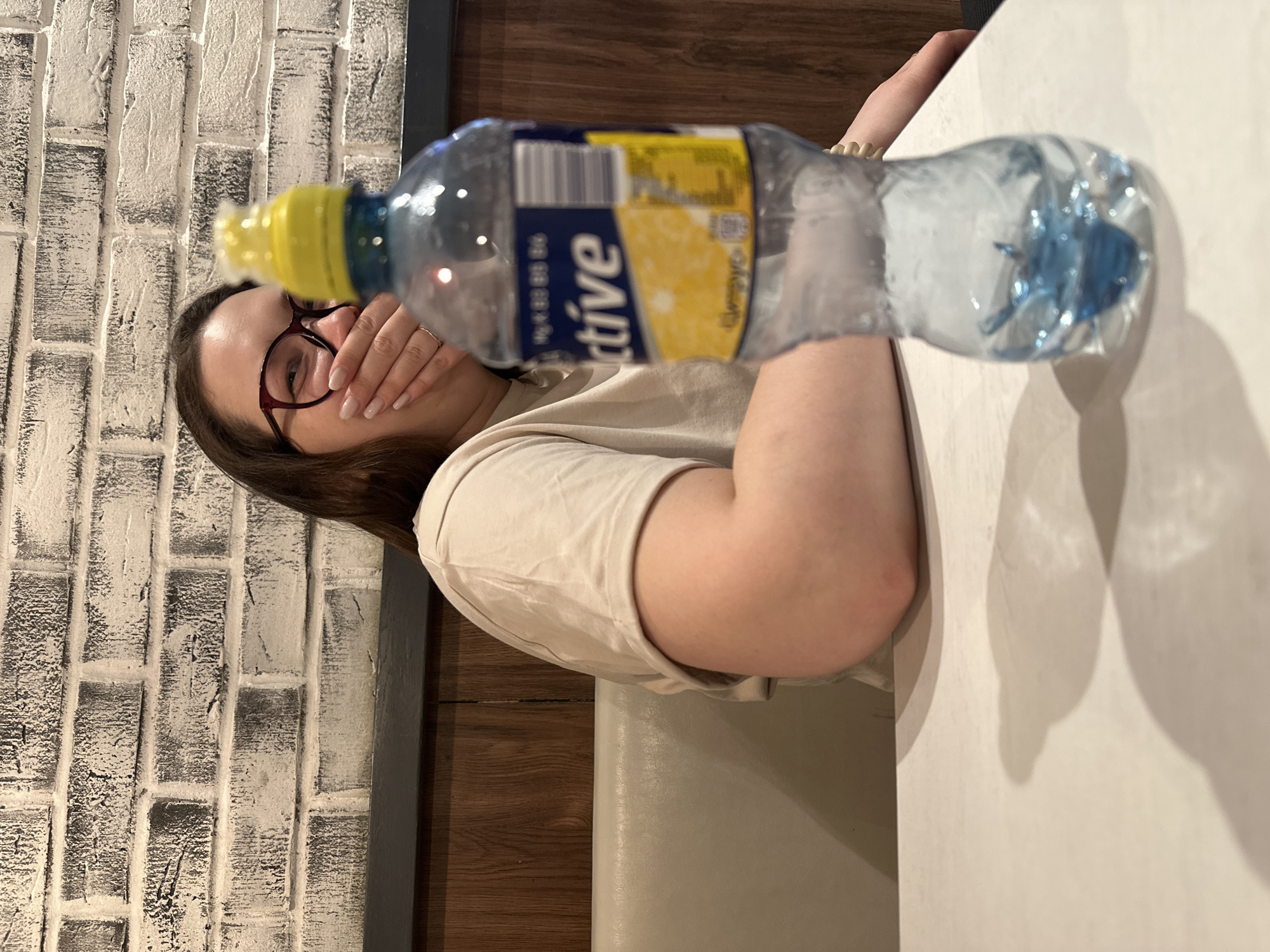 A woman with dark hair and red-framed glasses is partially obscured by a bottle of "Active" lemon-flavored water. She is seated at a white table, and her hand is near her face. The background features a white brick wall and dark brown wood paneling. The location is likely in Chaikovsky, Russia, based on the bottle's label.