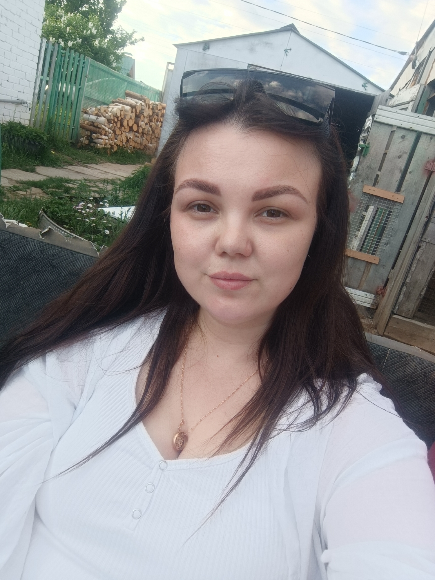 A medium shot captures a woman in Zav'yalovo, Russia, wearing a white, long-sleeved shirt with a buttoned neckline and a gold necklace with a locket. Dark sunglasses rest on top of her head. Behind her, a green wooden fence is visible, along with a stack of logs. Faded buildings are in the background. The sky appears overcast.