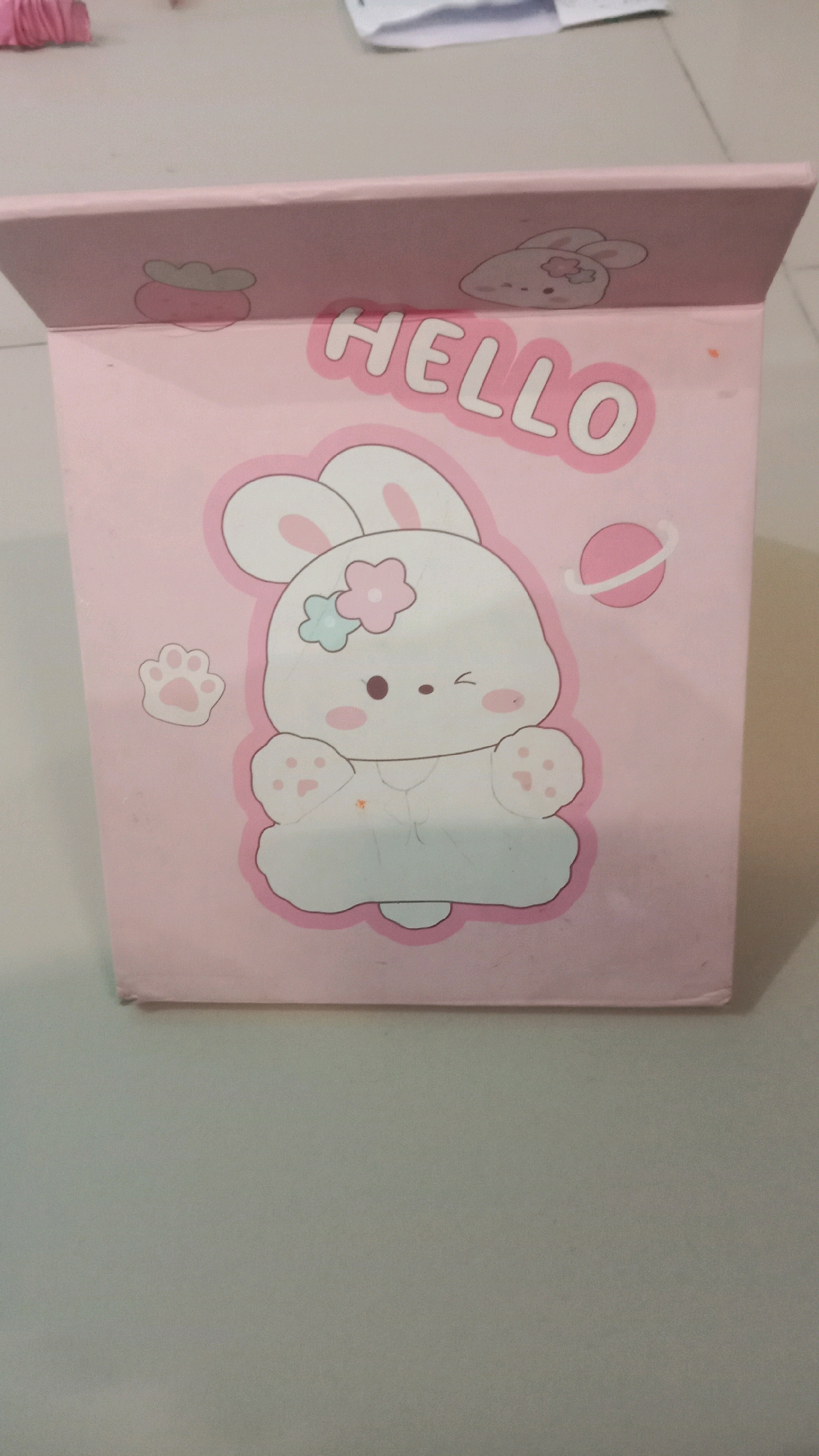 An overhead shot shows a pink, cartoon-themed storage box with a rabbit illustration and the word "HELLO" in white, puffy letters. The box is slightly tilted forward, resting on a neutral-colored, flat surface. The background is blurred, but appears to be a table. The image is not taken in Jalenjaya, Indonesia.