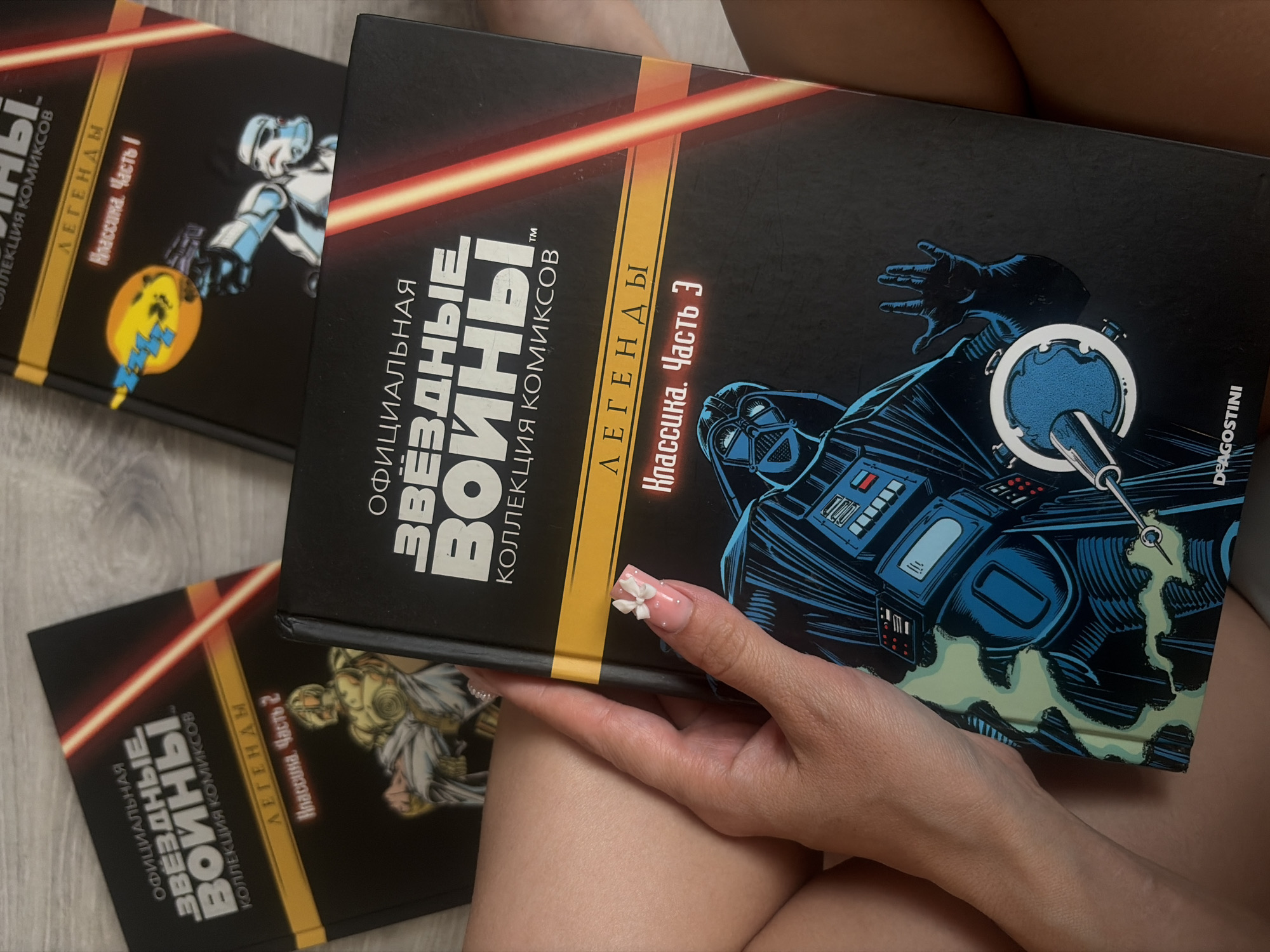 In an overhead shot, three Star Wars comic collections are arranged on a surface, with a person's hand and leg visible in the foreground. The top right comic features a stormtrooper aiming a weapon at a yellow circular object. The middle comic, held by the person's hand, displays Darth Vader on its cover and the text "ОФИЦИАЛЬНАЯ ЗВЕЗДНЫЕ ВОЙНЫ КОЛЛЕКЦИЯ КОМИКСОВ" (Official Star Wars Comic Collection) and "ЛЕГЕНДЫ Классика. Часть 3" (Legends Classic. Part 3). The person's hand has a pink ombre manicure with a small white bow on the ring finger. The background is blurred, suggesting an indoor setting. The lighting is soft and natural.