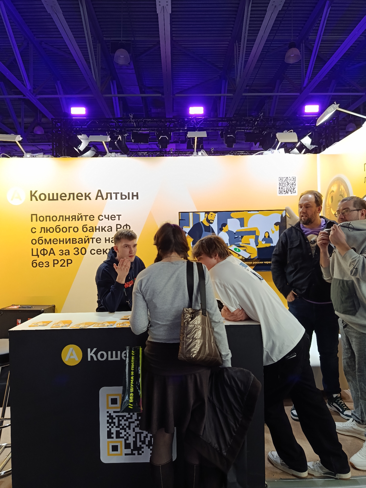 The image shows a booth for "Кошелек Алтын" (Wallet Altyn) in what appears to be an exhibition hall. The booth features a yellow and black design with the company's logo and a QR code. A young man is standing behind the counter, possibly assisting customers. Two people are standing at the counter, engaging with him. To the side, a group of three men are watching the interaction, with one holding a phone, possibly taking a picture or video. The booth's backdrop has the company's name, promotional text in Russian, and an illustration or screen display. The setting is indoors, with visible ceiling structures and lighting fixtures, giving the impression of a trade show or similar event.