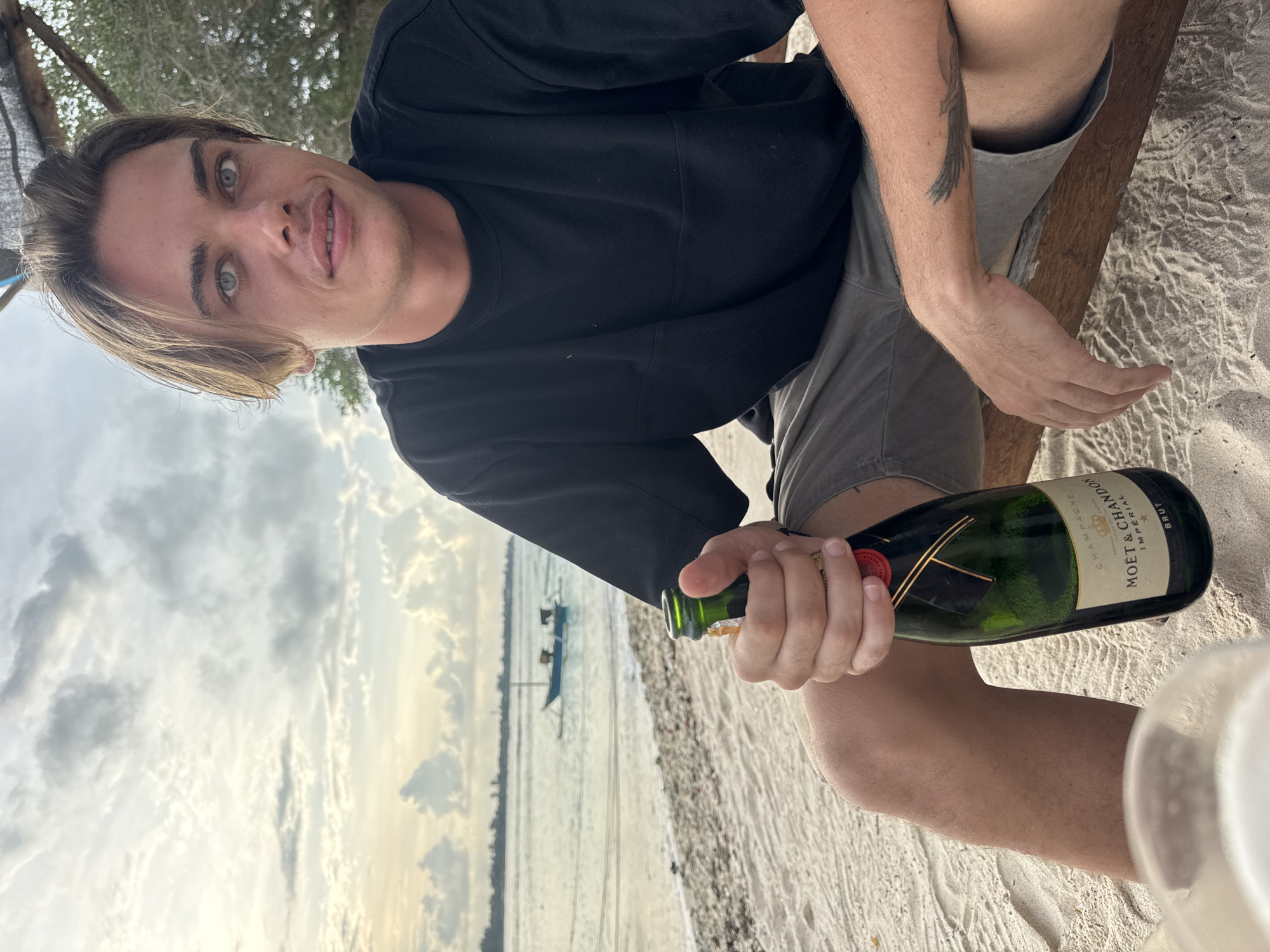 A man sits outdoors, holding a bottle of Moët & Chandon Impérial Brut champagne, at a beach in Unknown, Indonesia. The man has fair skin, light blonde hair, and grey eyes. He is wearing a black t-shirt and grey shorts. His left arm has a tattoo. In the background, the sea and the sky with patchy clouds are visible, with a small boat floating in the distance. The man is seated on a wooden structure on a sandy beach. The image is rotated counter-clockwise.