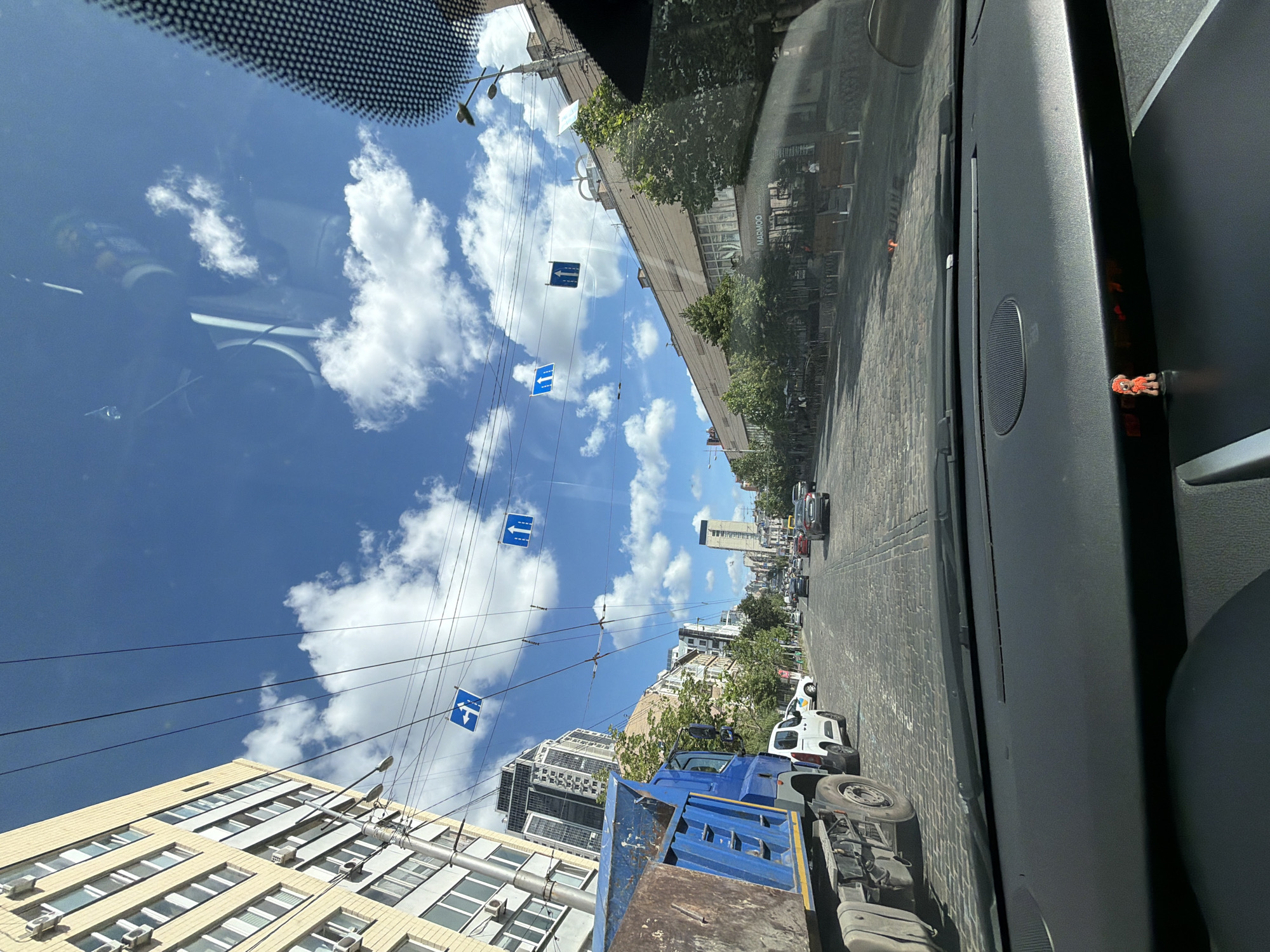 The image captures a view from inside a car, looking out through the windshield onto a street in Kyiv, Ukraine. The street is paved with cobblestones and lined with buildings and trees. Overhead, numerous power lines crisscross the sky, from which several blue traffic signs are suspended. These signs indicate traffic direction, with some showing arrows pointing straight or to the left, and others indicating a one-way street.
On the left side of the street, a large blue dump truck is parked. Further down the street, other vehicles, including cars and a white van, are visible, suggesting moderate traffic. Buildings of various architectural styles line both sides of the street, some appearing to be residential with multiple windows and air conditioning units. One building has a sign that reads "MARMOO".
The sky is blue with scattered white clouds, indicating a sunny day. The reflection on the windshield shows some interior elements of the car, including a dashboard with a small orange figurine and part of a seat. The overall scene depicts a typical urban street in Kyiv on a bright day.