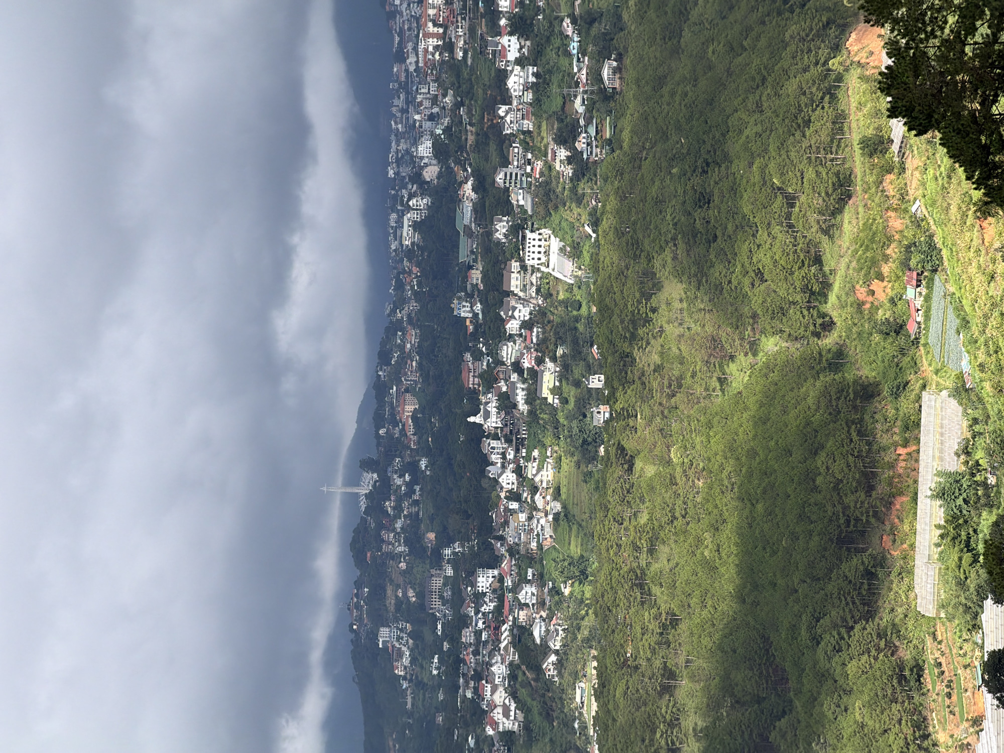 This is a high-angle shot of a densely populated hillside in Xuân Thành, Vietnam. The city is built on a steep incline, with houses and buildings clustered together. Lush green pine forests cover the lower slopes and extend up the mountainside. The sky is overcast with heavy gray clouds, casting a shadow over parts of the landscape. In the distance, a prominent white tower stands on a hilltop, and a complex of greenhouses is visible at the bottom of the frame. The architecture of the buildings in the city is varied, with many featuring white facades and red or dark-colored roofs, suggesting a mix of residential and commercial structures. The terrain is rugged, with winding roads and terraced areas interspersed among the trees and buildings.