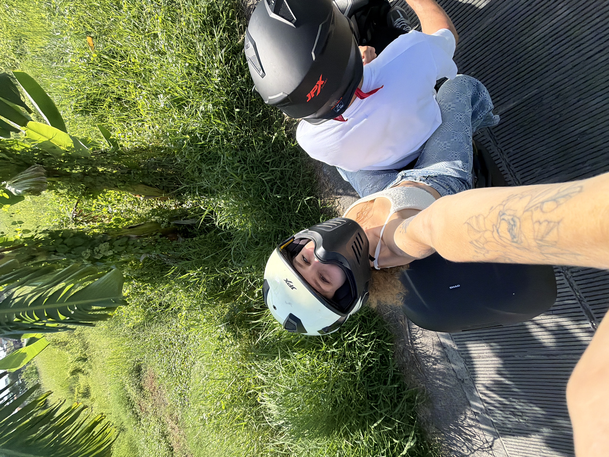 Two individuals wearing motorcycle helmets are on or near a motorcycle or scooter, which appears to be parked on a textured surface, possibly a paved area or walkway. Lush green grass and tropical plants are visible around the parked motorcycle. One individual is wearing a white top, patterned blue jeans, and has a tattoo on their arm. The other individual is wearing a white shirt. The location is North Kuta, Indonesia, based on the environmental context.