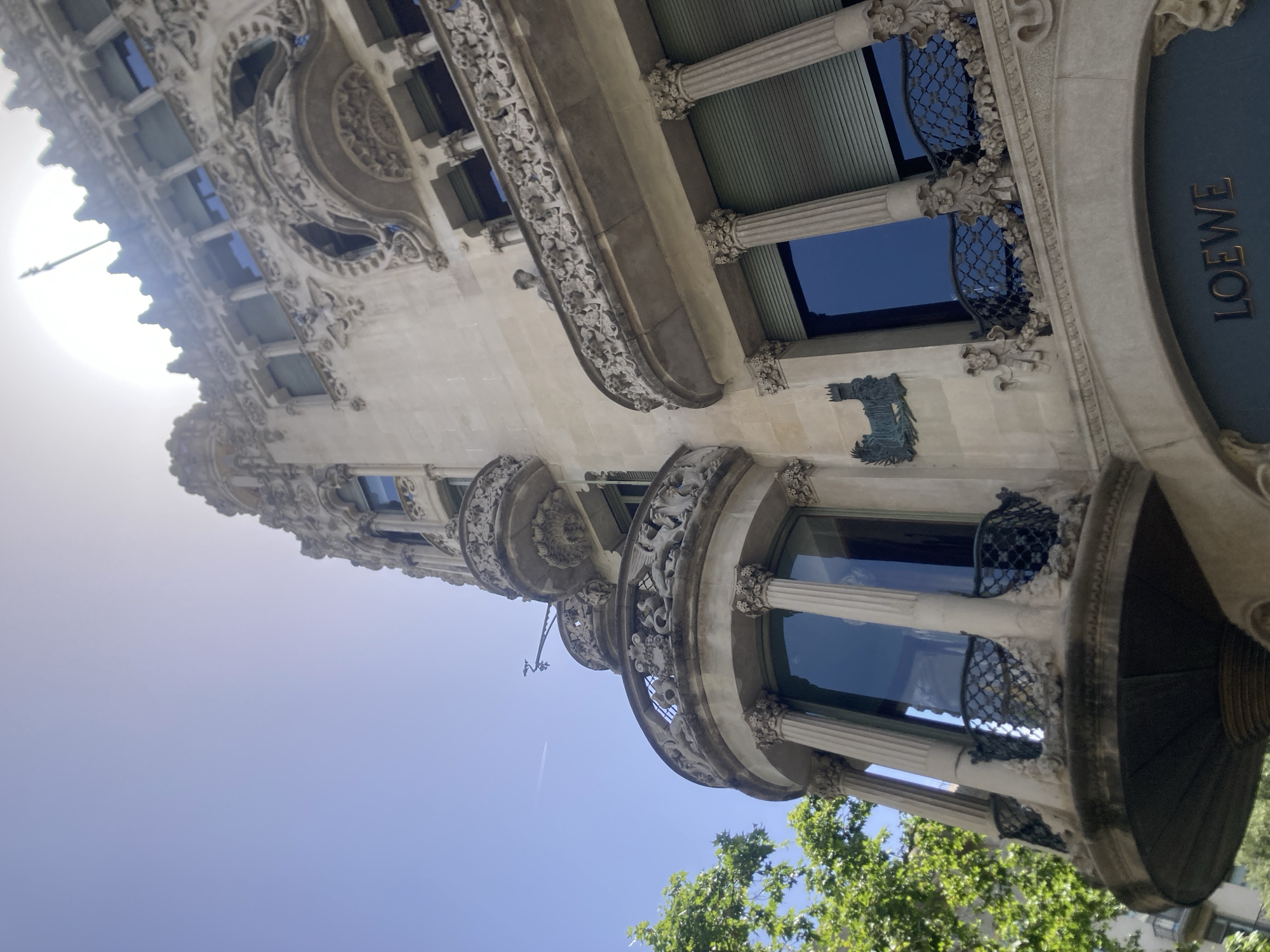 The image shows a close-up, low-angle view of an ornate building facade, possibly a historic structure, in Barcelona, Spain. The architecture is elaborate, featuring intricate stone carvings, decorative balconies with wrought-iron details, and fluted columns. The building's exterior is made of light-colored stone, with dark blue windows reflecting the sky. One section of the facade has a dark blue panel with the gold lettering "LOEWE" visible, indicating a luxury retail store.
The scene is set outdoors under a clear blue sky, suggesting a sunny day. The sun is visible in the upper left portion of the image, casting a bright glare. Green foliage from trees is visible at the bottom right, adding a natural element to the urban setting. A faint contrail from an airplane can be seen in the sky. The overall impression is one of architectural grandeur and a bright, pleasant day. The focus is on the detailed craftsmanship of the building's facade and the juxtaposition of historical architecture with modern retail.
