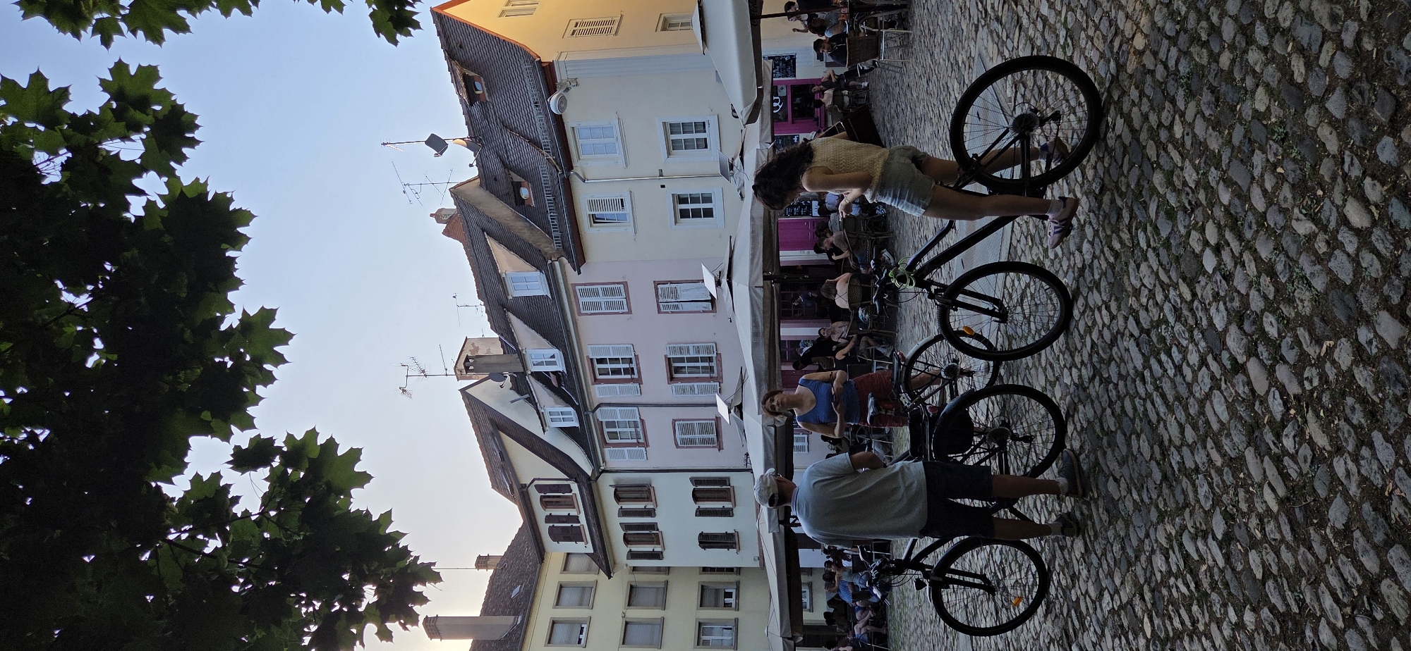 In an outdoor European town square, illuminated by the warm glow of the late afternoon sun, two individuals stand by their bicycles on a cobblestone ground. The scene is framed by the dark silhouette of leafy tree branches overhead. In the background, a row of buildings with multi-colored facades, featuring traditional shutters and antennas on their roofs, form a picturesque backdrop. Large white umbrellas and tables suggest a lively outdoor cafe or restaurant area, where numerous patrons are seated. The man, wearing a light blue t-shirt, shorts, and a baseball cap, faces the woman. The woman, in a blue tank top and red shorts, is looking down at her phone while leaning against her bicycle. Another bicycle is positioned to the left of the man. The overall atmosphere is one of relaxed urban leisure.