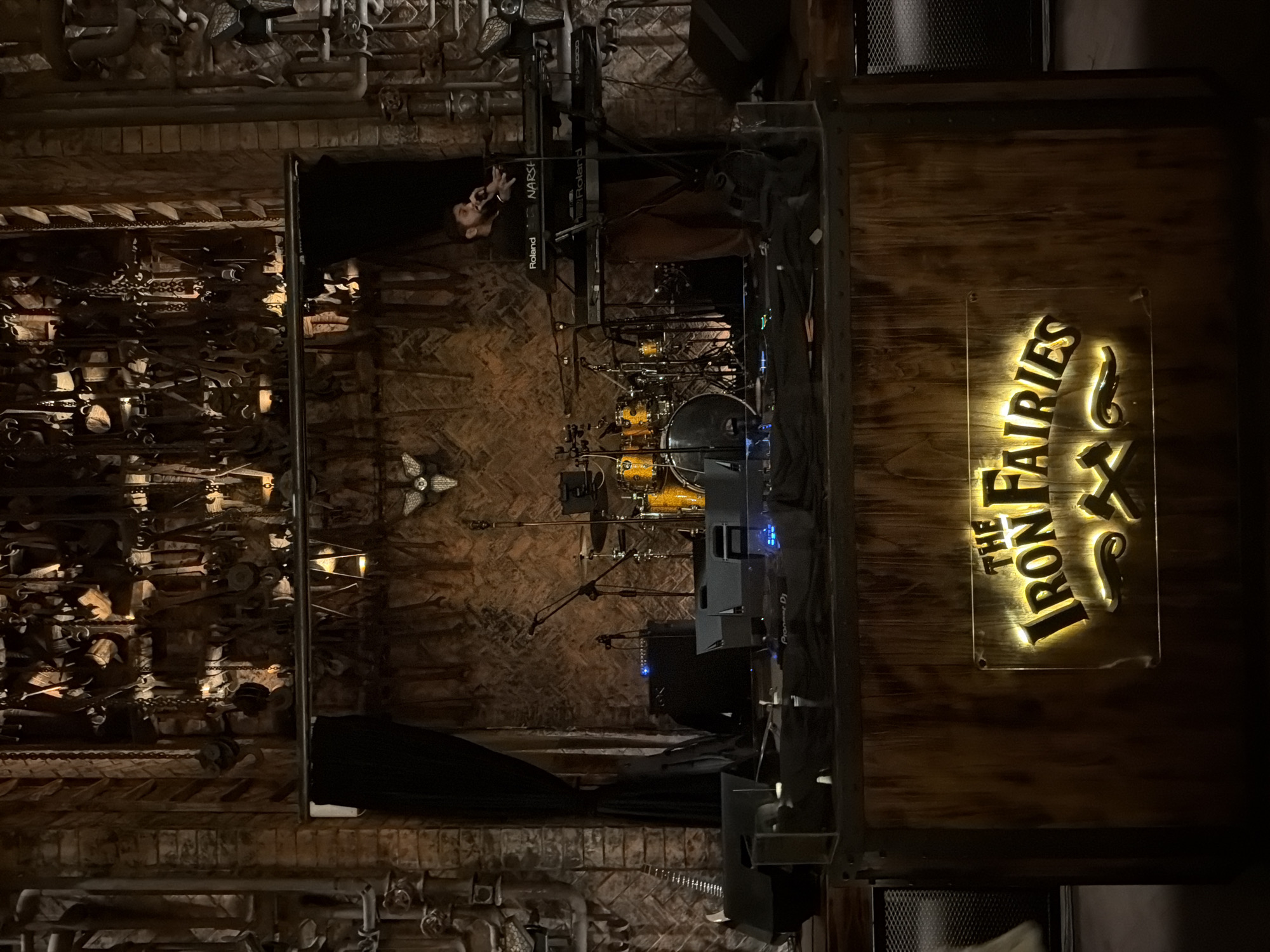 The image depicts the stage of a dimly lit indoor music venue, identified as "The Iron Fairies" in North Kuta, Indonesia. The stage is set with musical equipment, including a drum kit, a keyboard with a musician playing it, microphones, and amplifiers. The backdrop is a textured wall with a display of various tools and chains hanging from above. The overall aesthetic is rustic and industrial, with exposed brickwork and wooden elements, including the front of the stage which features a glowing sign with the venue's name and a stylized logo. A single musician, dressed in dark clothing, is actively playing the keyboard and singing into a microphone. The lighting is atmospheric, with spotlights focused on the stage and the venue's signage, creating a dramatic effect.