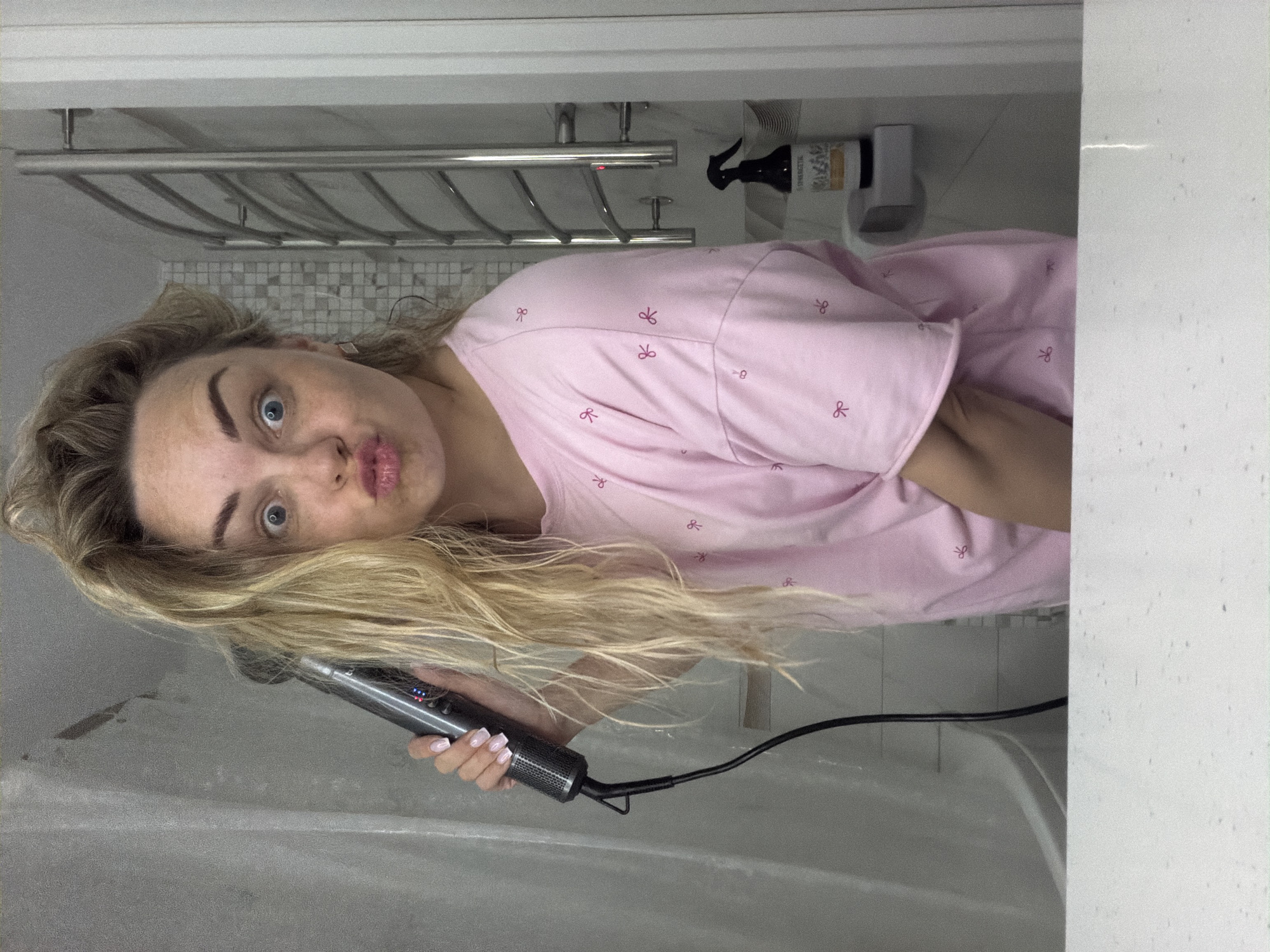 A woman with blonde hair is in a bathroom, holding a hair styling tool. She is wearing a pink shirt with a bow pattern. The bathroom contains a towel rack, a cleaning spray, and a tiled wall. The location is not discernible as Votkinsk, Russia.