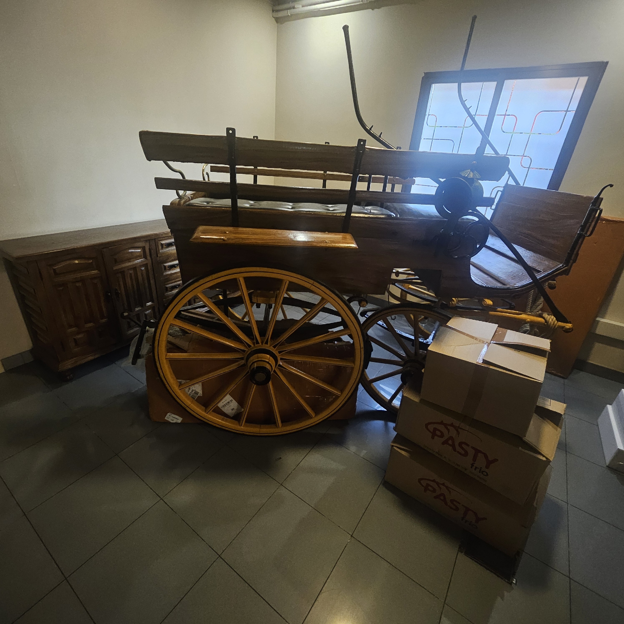The image depicts an indoor scene, likely a storage area or a room undergoing setup. The primary subject is a wooden carriage, possibly an antique or replica, with large yellow spoked wheels. The carriage has a bench seat and appears to be made of dark wood with metal accents. To the left of the carriage is a dark wooden cabinet with multiple drawers and doors, exhibiting ornate carvings. In the foreground and to the right of the carriage, several cardboard boxes are stacked. The boxes are brown and some feature a red logo that reads "PASTY trio". The floor is tiled with large, dark gray square tiles. In the background, a large window or glass panel with a geometric design is visible, leaning against the wall. The walls are painted a light, neutral color, and the ceiling shows some light fixtures and ventilation elements. The lighting in the scene is somewhat dim, casting shadows. There are no people visible in the image. The context provided states the location as Almadrones, Spain, suggesting this is a scene within that municipality.