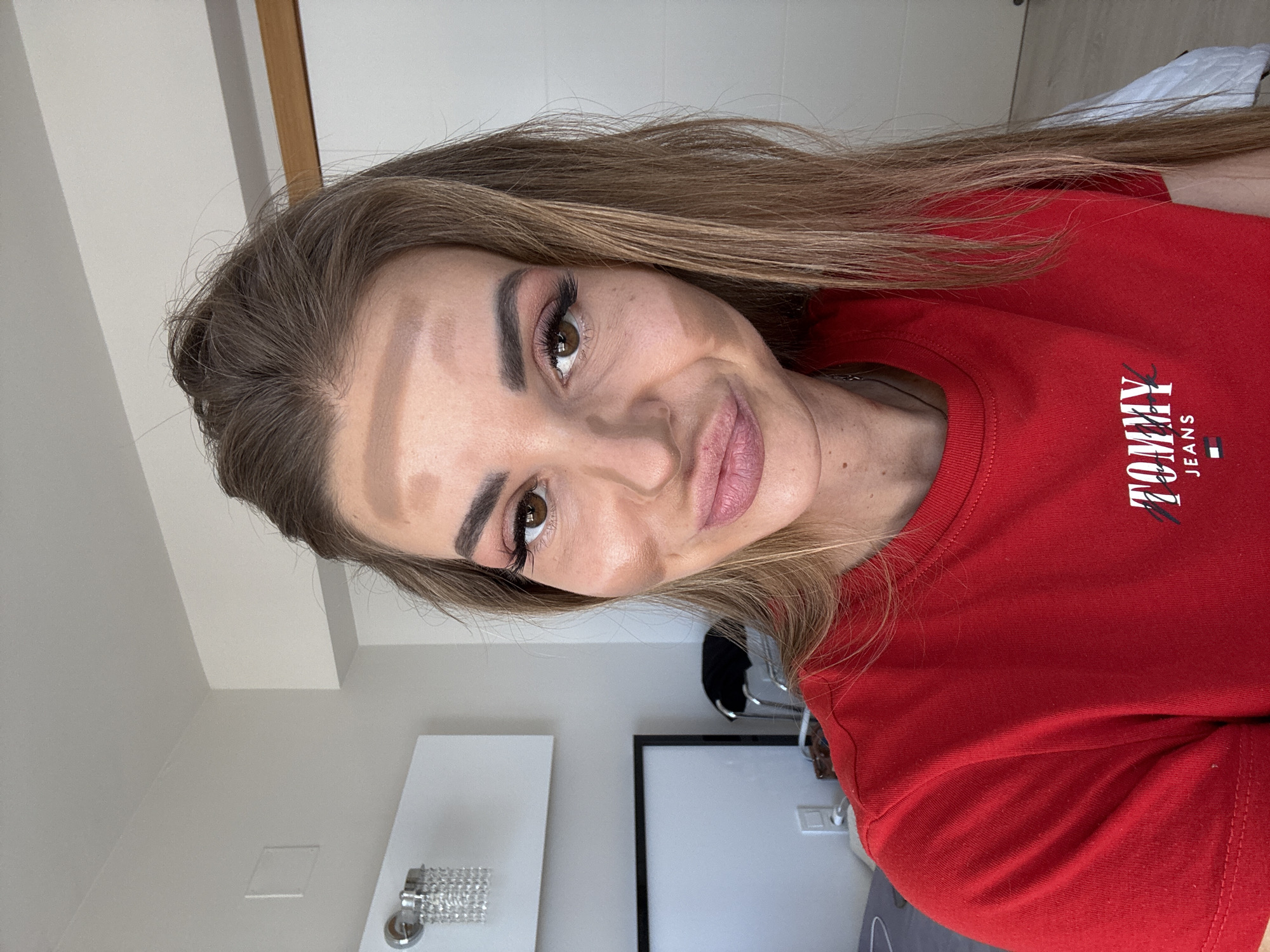 A woman is looking at the camera while wearing a red Tommy Jeans t-shirt. Her hair is long and brown. The background shows a white wall, a chandelier, a black framed object, and a chair. The location information is not available in the picture.