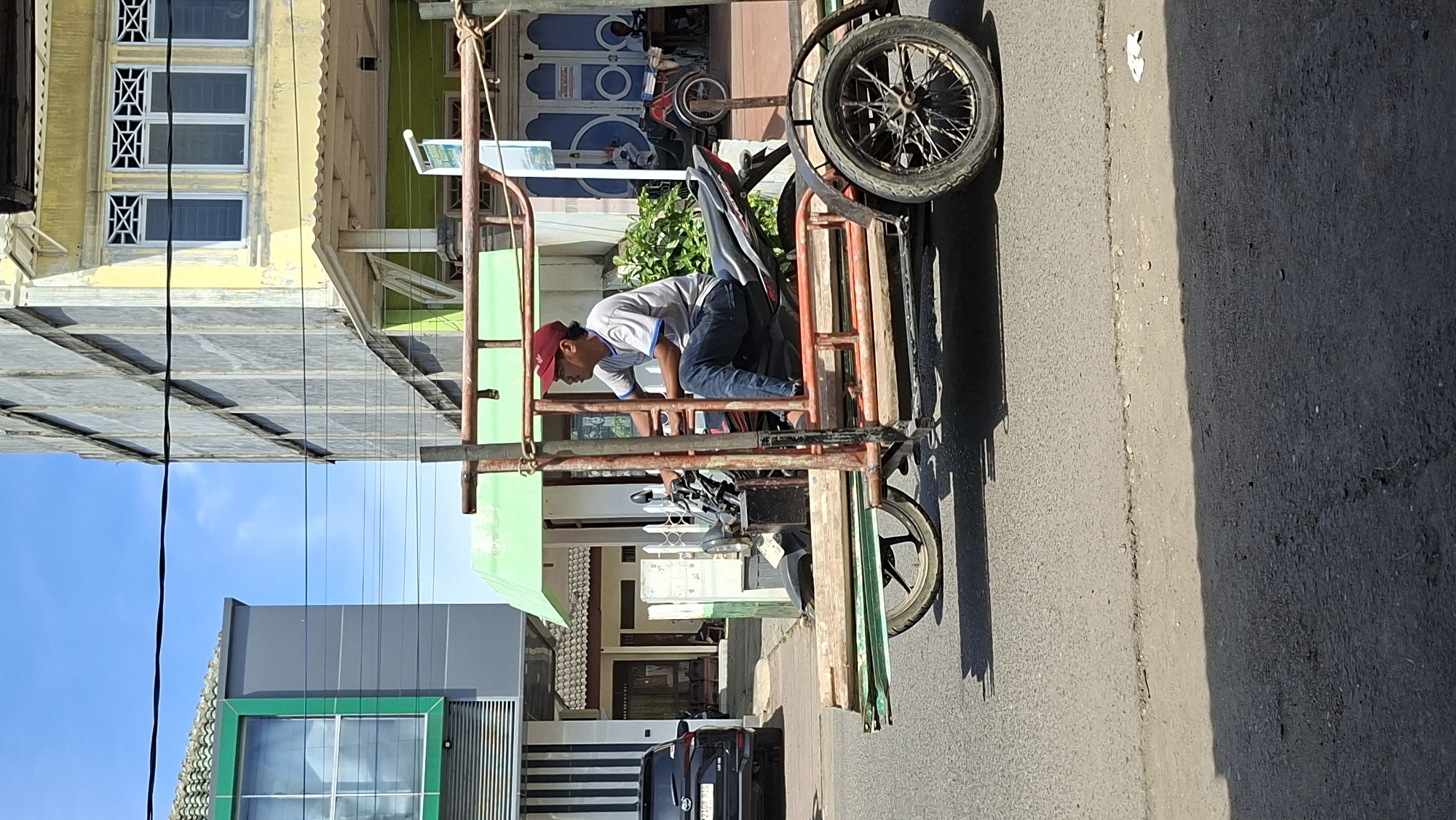 An outdoor, eye-level, medium shot captures a man riding a three-wheeled motorcycle with a cargo trailer in Lhokseumawe, Indonesia. The man, wearing a red baseball cap, a grey t-shirt, and dark blue jeans, sits on the motorcycle seat, looking to the left. The motorcycle is attached to a trailer constructed of metal tubing and wooden planks, carrying several pieces of lumber. The trailer has two spoked wheels at the rear and one at the front, partially obscured by the motorcycle. The scene is set on a paved road with a visible curb. In the background, buildings line the street. On the left, a modern building with a green trim and a glass facade is visible. Adjacent to it is a building with a yellow facade, featuring decorative windows, and a green awning. The sky above is clear and blue.
