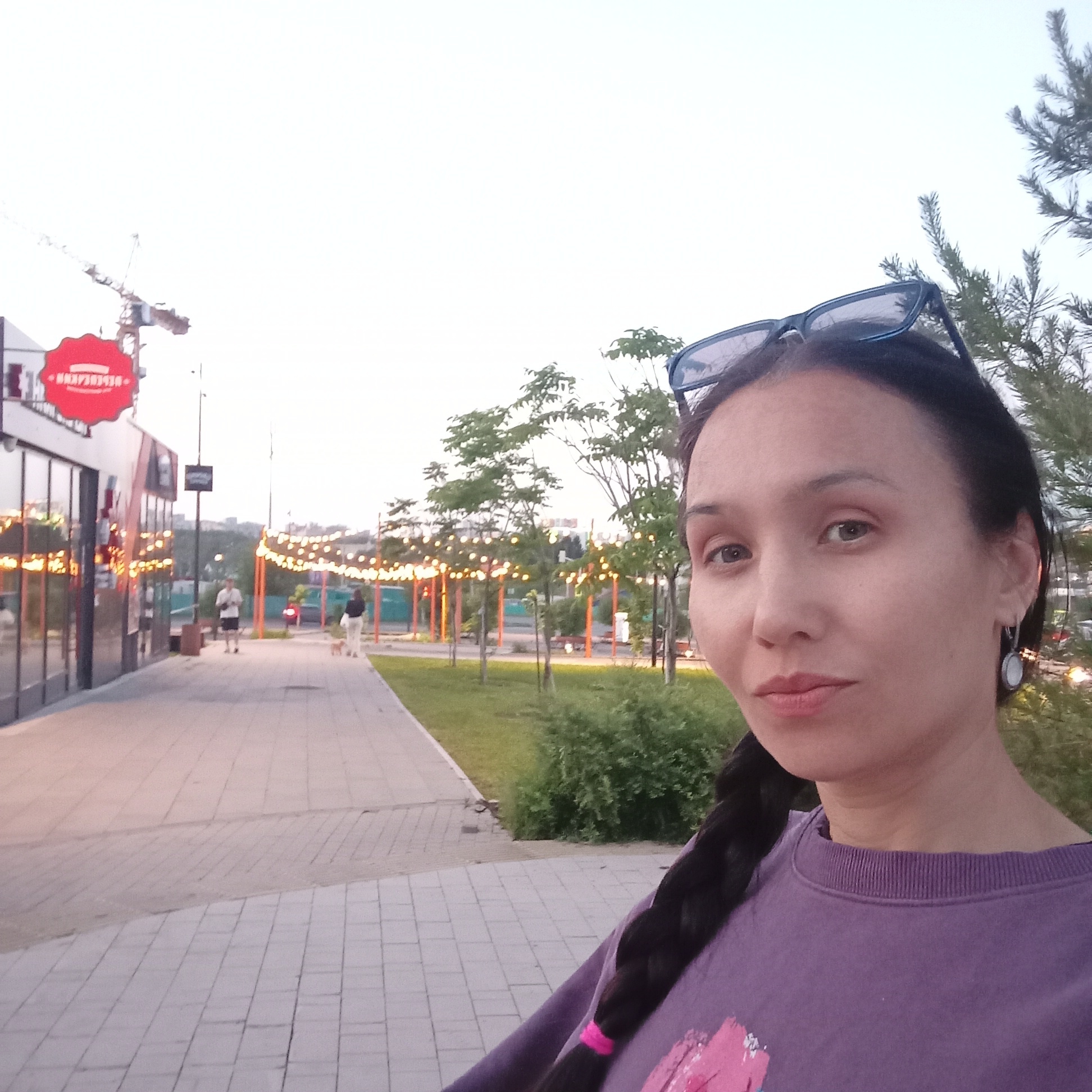The image displays a woman taking a selfie at twilight in Pervomaysky, Russia. The woman is positioned in the right foreground, depicted from the chest up. She has dark hair styled in a long braid, secured with a pink tie visible over her left shoulder. Dark sunglasses rest on top of her head. She wears a purple long-sleeved sweatshirt adorned with a pink and white graphic design on her left chest area. A circular white and silver earring hangs from her right ear.
In the midground, a paved pedestrian walkway, constructed from light-colored pavers, extends from the left towards the background. To the right of the walkway lies a grassy area interspersed with young trees and bushes. String lights are strung between orange support poles, forming an illuminated canopy over a portion of the path and green space further down. Two individuals are visible on the walkway in the distance: one person walks alone, and another walks a small, light-colored dog on a leash. On the left side of the walkway, a modern building with large glass window panels reflects ambient light. A prominent red, scalloped-edge sign with white Cyrillic text is affixed to the building's exterior. In the background, a construction crane is silhouetted against the light, overcast sky, rising above the distant cityscape.