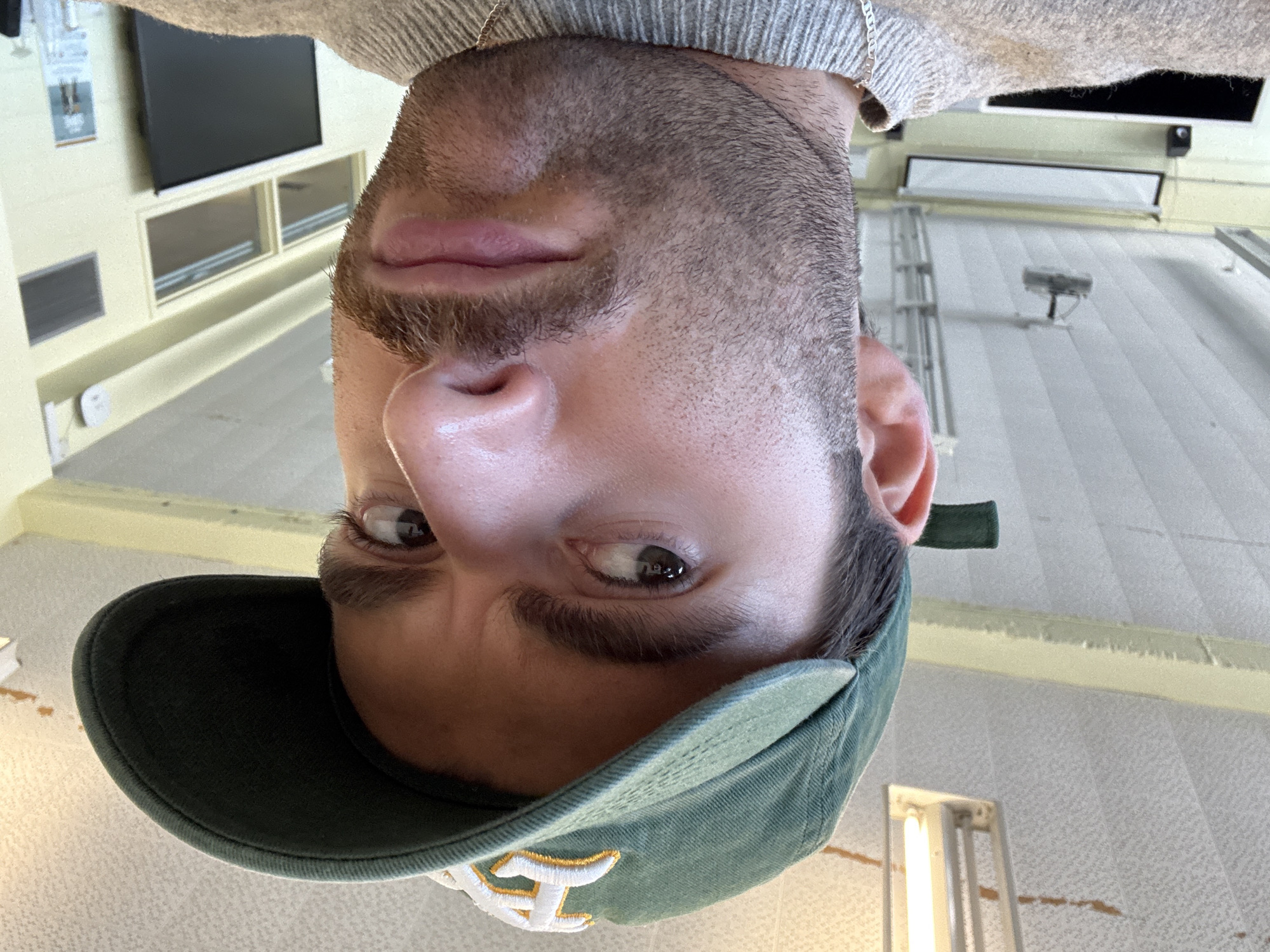 The image, captured from a low, upward angle and presented upside down, focuses on the face of a young man, likely in his 20s or 30s. He has fair skin, short dark hair, and a short beard or heavy stubble. He is wearing a light grey knitted sweater and a dark green baseball cap with a stylized yellow or off-white letter, possibly an 'A' or 'M', embroidered on the front. A thin silver chain is visible around his neck. His brown eyes are open and directed towards the viewer with a neutral, slightly watchful expression, and his lips are slightly parted.
The background indicates an indoor setting, likely a commercial or institutional building, possibly in Montreal, Canada, with high ceilings. To the left side (when the image is conceptually righted), a light-colored wall features a black flat-screen television mounted high up, with two rectangular window-like panels or displays below it, and a dark grey rectangular vent further down. To the right, the ceiling or upper wall has a textured surface, possibly acoustic tiles or wallpaper, with visible seams. Fluorescent light fixtures are attached to the ceiling, and in the distance, a ladder or scaffolding structure is visible, along with a small, dark device, possibly a security camera, mounted on the ceiling. The floor, out of focus, appears to be a light-colored carpet. The lighting suggests it is daytime. The man appears to be taking a selfie or posing for a photo from this unusual perspective.