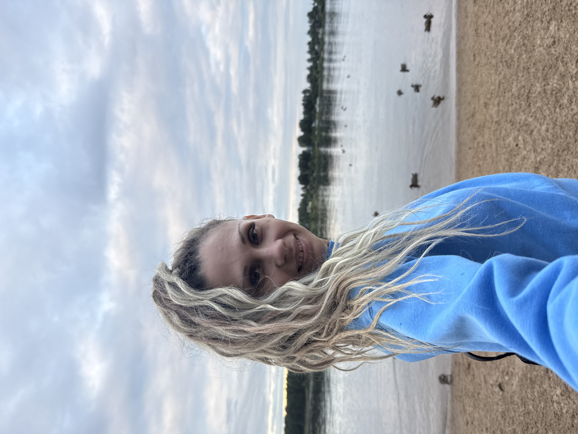 A medium shot captures a woman with long, blonde, dreadlocked hair pulled into a high ponytail, facing the camera from a slightly angled profile. She is wearing a bright blue sweatshirt. The background depicts a body of water, likely a lake, with a treeline visible on the far shore. Several ducks are swimming in the water. The foreground consists of a sandy shore. The sky above is overcast with clouds. The scene appears to be a casual outdoor setting in Rusanovo, Russia.
