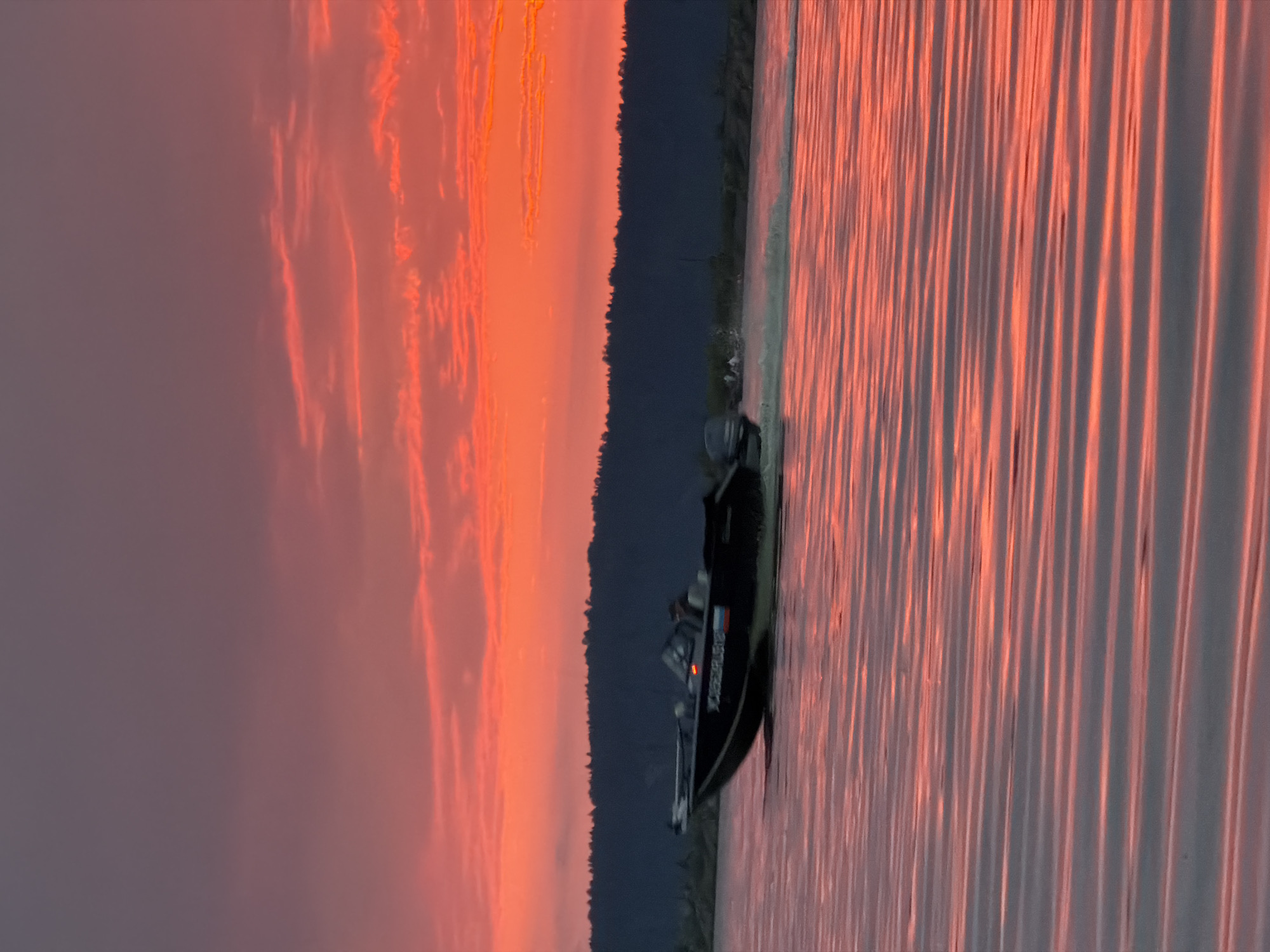 An image captures a black motorboat moving at speed across a body of water, leaving a visible wake. The boat is dark in color with some decals or writing visible on its side. Two individuals are seated within the boat. The water surface is rippled and reflects the vibrant hues of the sky, which is dominated by a dramatic sunset. The sky transitions from a deep pink and orange near the horizon to a muted purple-grey at the top. Behind the water, a dark, dense line of trees forms a treeline, suggesting a forested area. The overall scene is set during twilight, illuminated by the colorful sky. The location is identified as the Kama River in Chaikovsky, Russia.
