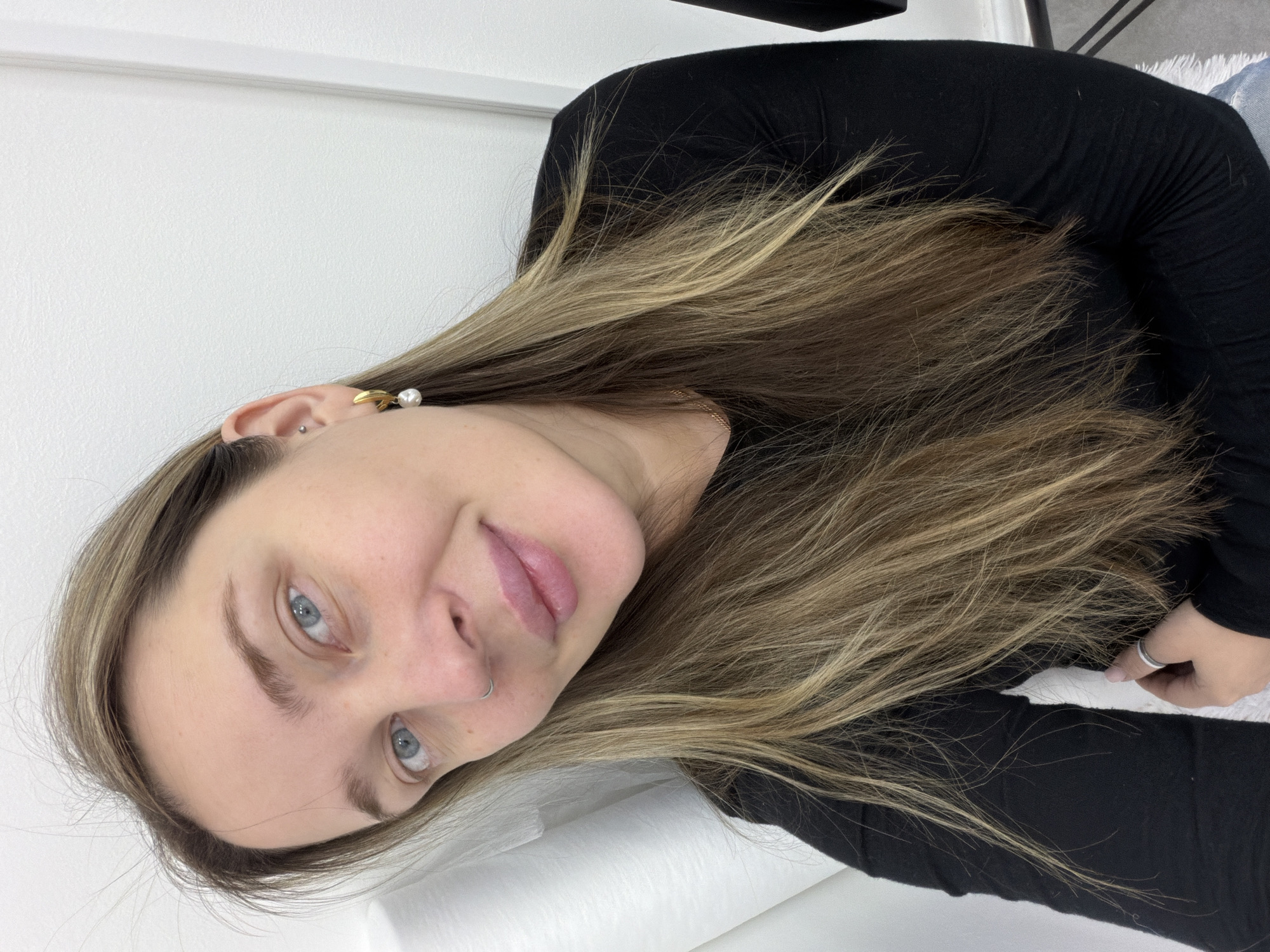 A woman with long, highlighted brown hair is lying on a white surface, possibly a bed or examining table, and looking directly at the camera. She has blue eyes, fair skin, a nose piercing, and gold and pearl earrings in her left ear. She is wearing a black long-sleeved shirt. Her hand, with a silver ring, is visible near the edge of the frame. The background consists of a white wall. The image provides no evidence to suggest the location is Moscow, Russia.