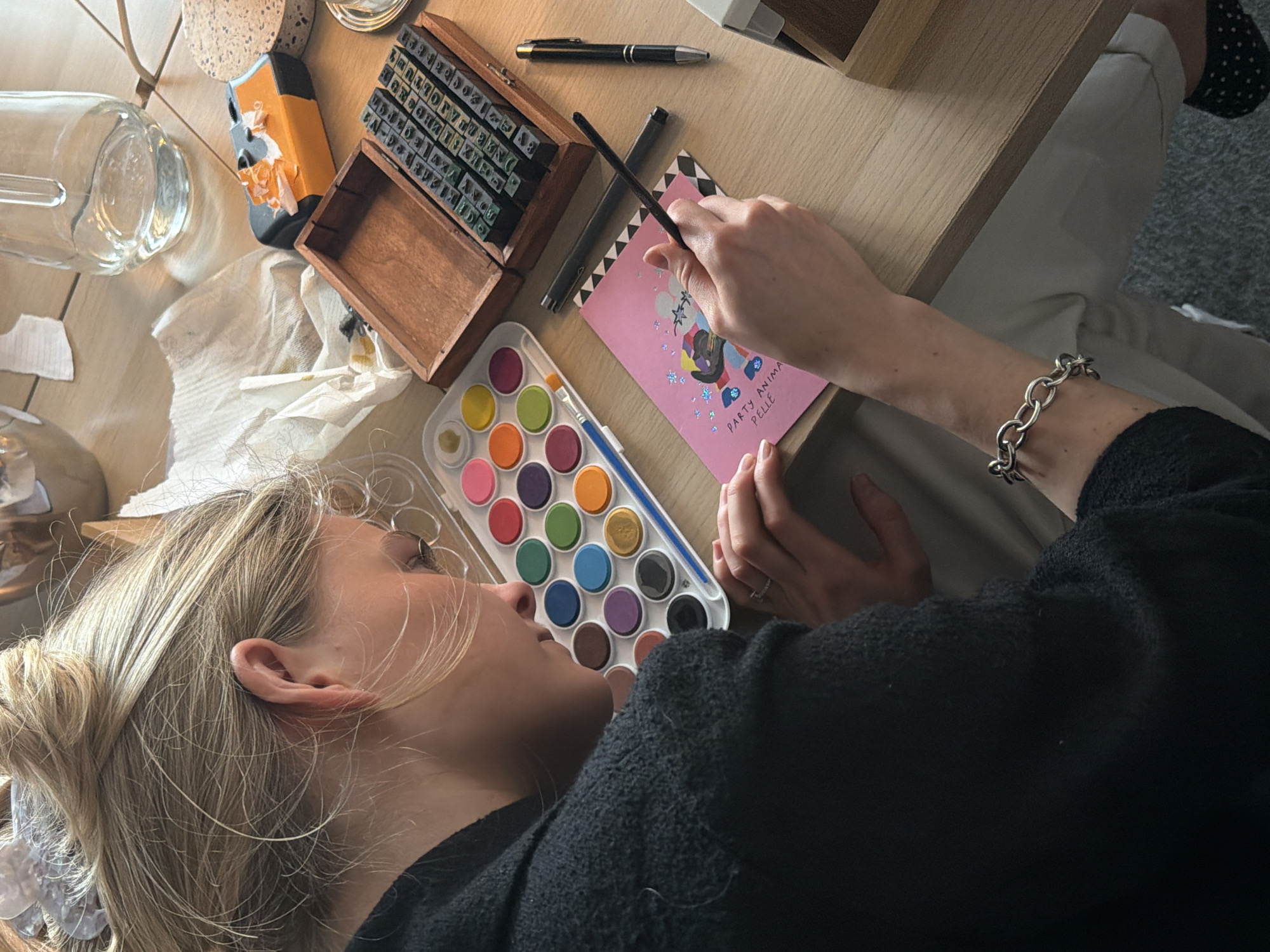 A young woman with blonde hair, styled in a messy bun, is partially visible in this top-down shot, engaged in an art activity at a light-colored wooden table in an indoor setting. She is wearing a dark, possibly knitted, long-sleeved top, and has a silver chain-link bracelet on her left wrist, along with a ring on her left hand.
Her hands are positioned over a pink card or piece of paper, which she is actively decorating with a paintbrush. The card features a colorful illustration, possibly of an animal character with balloons, and contains partially visible text that reads "PARTY ANIMAL PELLE" or "PARTY ANIMAL PAL." Next to her, a white plastic watercolor set with numerous colorful paint pans and a blue-handled paintbrush is open.
Other items on the table include a wooden box filled with a set of rubber letter stamps, two pens (one black with a silver clip), a clear glass bottle or jar, a crumpled piece of white paper or tissue, and a light-colored coaster. The woman appears focused on her creative task. The lighting suggests it is daytime. A glimpse of a foot in a dark, polka-dotted slipper and a light-colored carpet is visible in the background. This scene depicts a calm, creative moment, likely taking place in a home or studio in London, United Kingdom.