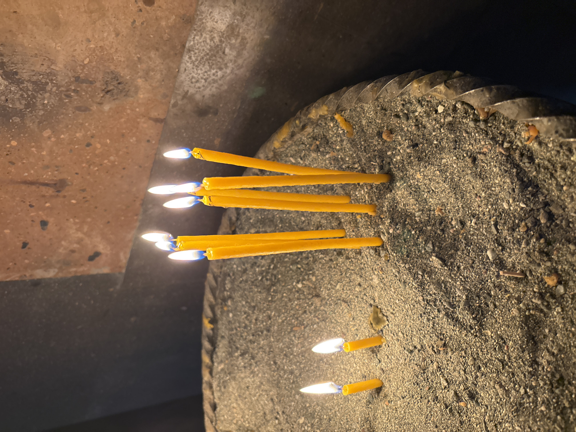 The image captures a close-up view of multiple yellow taper candles, several of which are actively burning, casting a warm glow. These candles are embedded in a substantial bed of dark sand or fine gravel contained within a large, shallow metal tray. The tray features a distinctive, possibly spiraled or braided, metallic rim that runs along its visible edge.
In the mid-ground, a cluster of longer candles stands upright, their wicks ablaze with bright, blue-tinted flames. Further back in the tray, two shorter, almost fully consumed candles are also lit. Melted wax residue is visible among the sandy bedding. The granular texture of the sand and gravel, composed of various particle sizes and shades of grey and brown, is clearly visible.
The background consists of a textured, dark brownish-grey surface, likely a wall or structural element, appearing rough and possibly made of stone or concrete. The scene is dimly lit, with the candle flames serving as the primary light source, creating pockets of illumination against the darker surroundings.
No people are visible in the image. The arrangement of candles in a sand-filled tray is a common practice in many Christian traditions, particularly in Orthodox churches and monasteries, where candles are lit as offerings, prayers, or acts of remembrance. Given the provided location context of Vagharshapat, Armenia, this scene most likely depicts a moment of worship or spiritual reflection within a church or monastic complex. The atmosphere conveyed is one of solemnity and quiet reverence. No text is visible in the image.