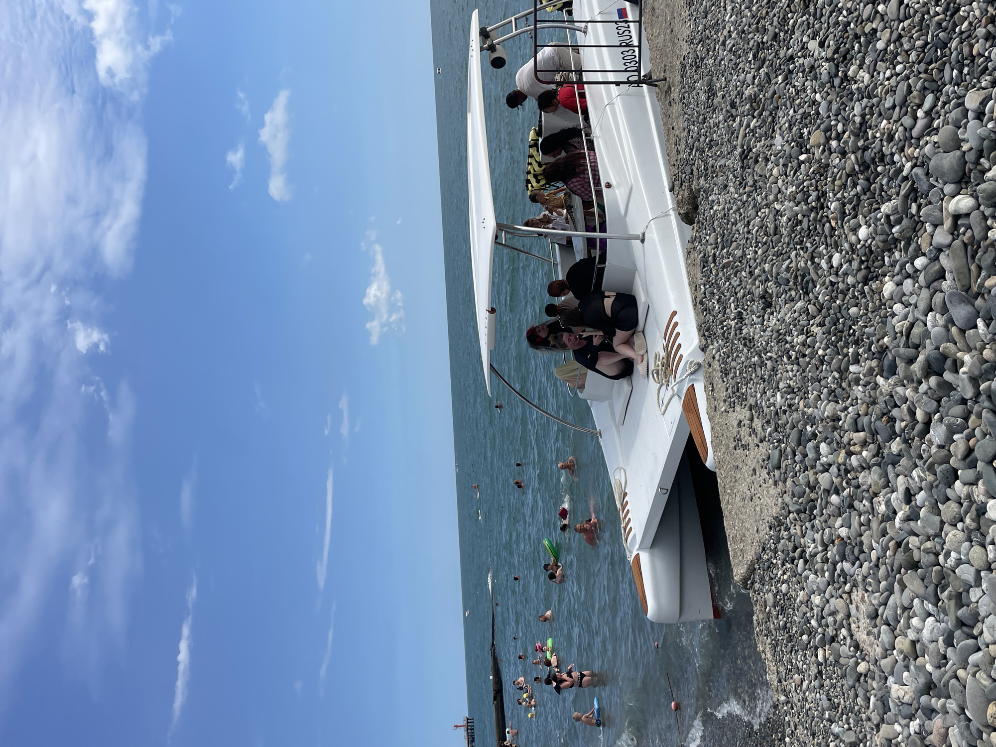 A white catamaran-style boat is moored on a pebbled beach, with its bow partially submerged in the dark blue water of the Black Sea. Several people are seated under the boat's canopy, while others are visible in the water, swimming and floating on inflatable rings. In the background, a red lighthouse stands on a pier, and the horizon is dotted with other small boats. The sky is a clear blue with scattered white clouds. The registration number on the boat reads "AO 0303 RUS22." The scene appears to be a leisure activity on a sunny day at a beach in Sochi, Russia, likely a popular tourist destination given the presence of the boat tour and numerous swimmers.
