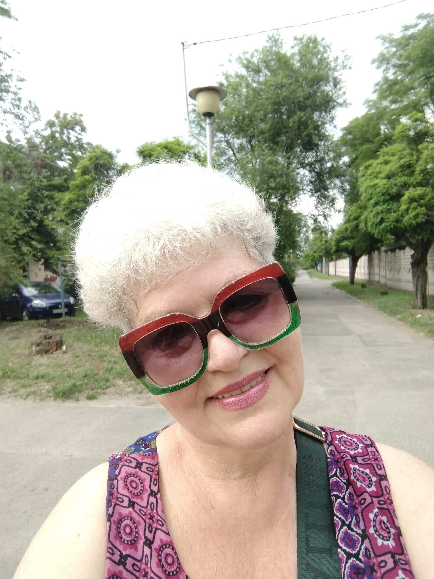 The image is a close-up, high-angle selfie of an older woman, presumably taken in Horishni Plavni, Ukraine. The woman has short, curly white hair and wears oversized sunglasses with thick frames that are red-brown at the top, black in the middle, and glittery green at the bottom. She has pink lipstick and a gentle smile. Her attire includes a sleeveless top or dress with a vibrant pattern featuring pink, purple, blue, and black geometric or floral motifs. A dark green strap from a bag is visible over her right shoulder.
The background shows an outdoor setting with a paved pathway extending into the distance on the right side of the frame. This path is bordered by a light-colored concrete block fence and a line of green trees. To the woman's left, a grassy area with a tree stump is visible, and further back, a blue car is partially parked. In the upper part of the image, a street light with a light-colored top is present, and numerous green trees fill the remaining background against an overcast, bright sky.