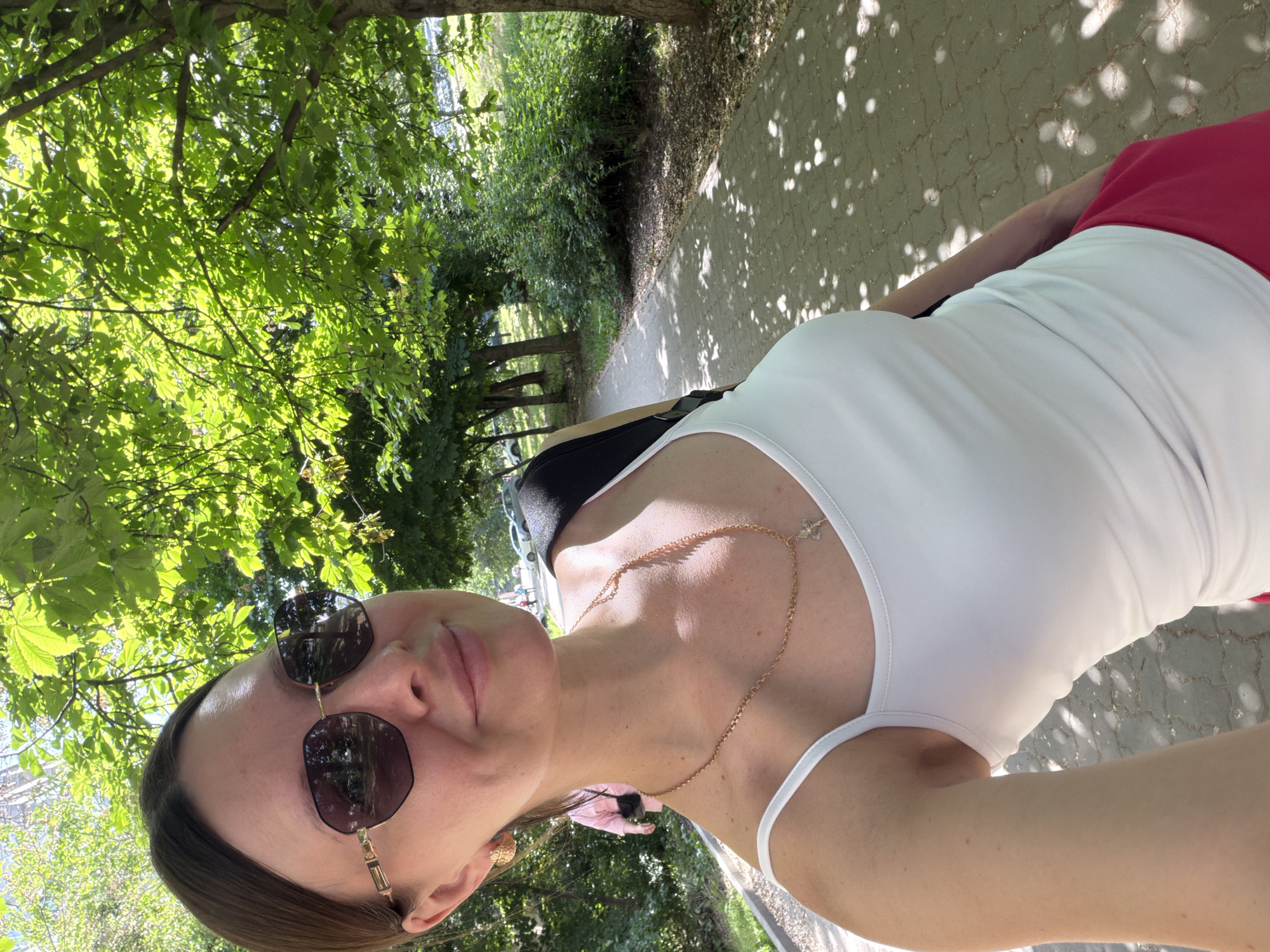 A woman is taking a selfie outdoors, likely on a path in a park or green space. She is wearing sunglasses, a white tank top, a gold necklace, and a red garment. The setting is a sunny day, with trees providing shade. The ground appears to be paved with dappled shadows. The location could be Rostov-on-Don, Russia, given the instructions.